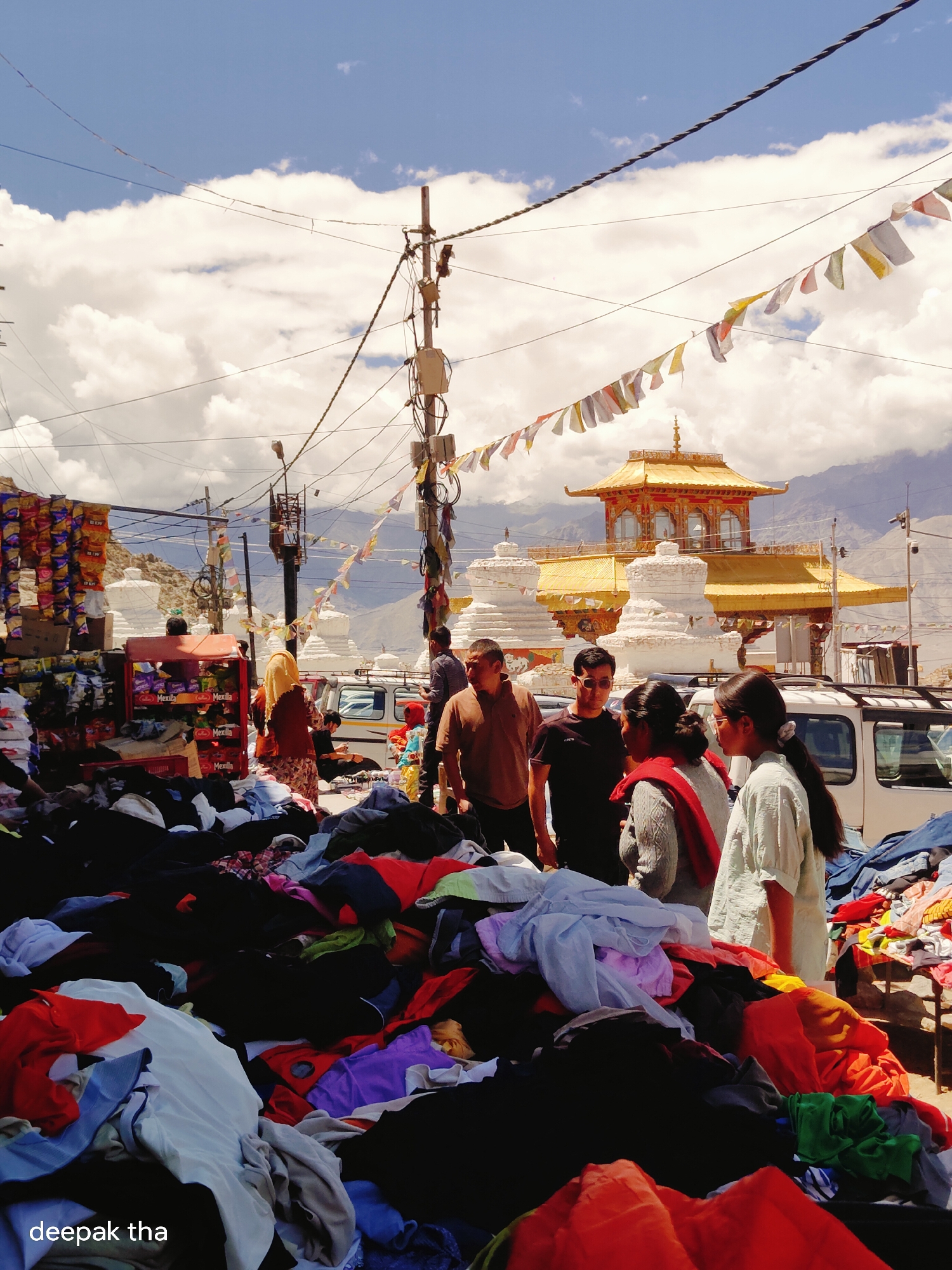This is a scene of an outdoor market in Leh, India, with a golden-roofed temple and stupas in the background. A street vendor displays stacks of clothing in the foreground. Several people are browsing the merchandise and walking around. The sky is blue with fluffy white clouds, and there are mountains in the distance. Prayer flags are strung across wires above the market. A power pole with many wires is visible.