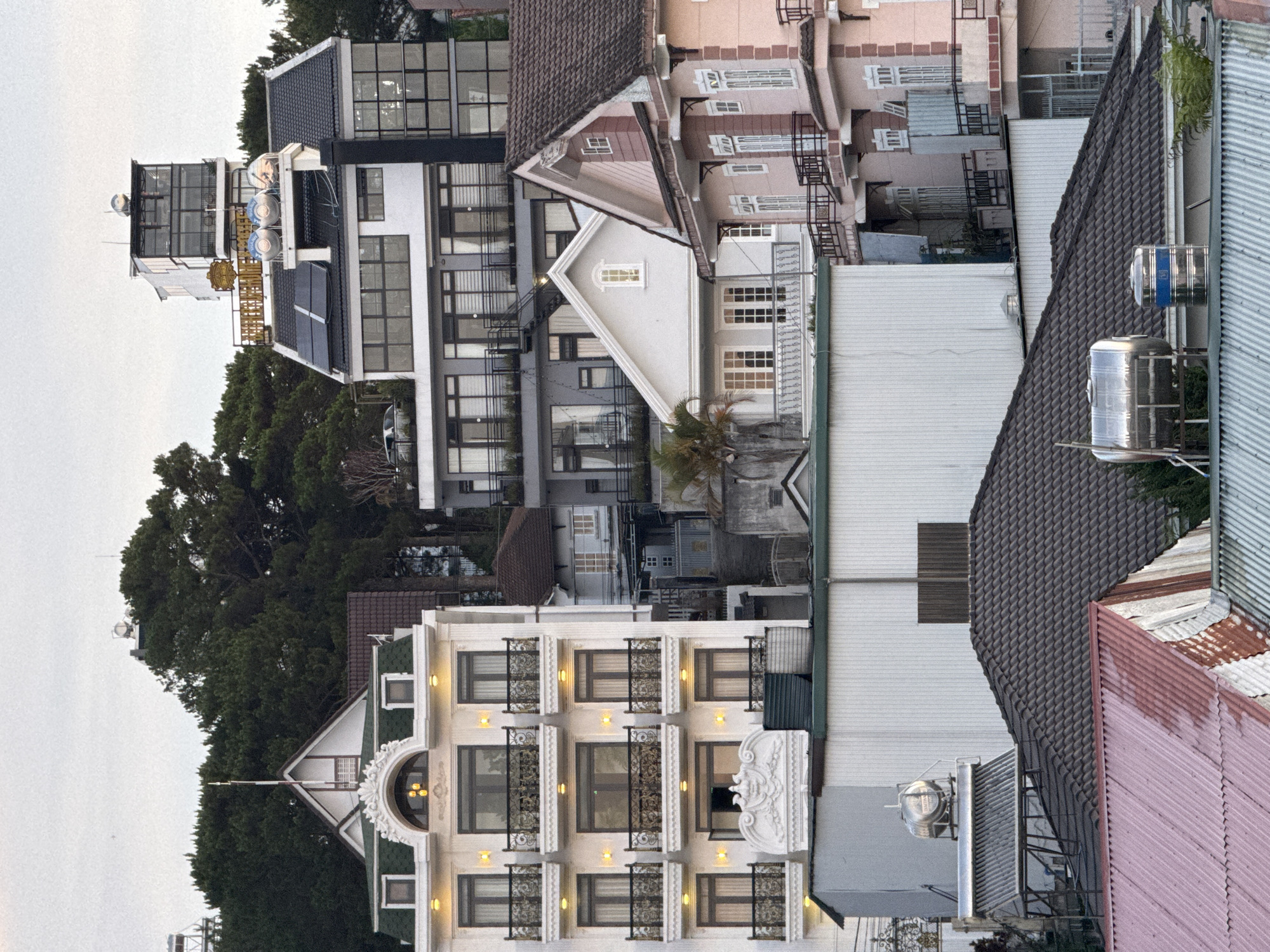 The image shows a dense, urban scene in the city of Lâm Viên - Đà Lạt, Vietnam. There is a mix of architectural styles with tall buildings, each with varying colors, materials, and shapes. A tall building with a predominantly white facade, multiple windows, and balconies with intricate iron railings dominates the lower center of the frame. Adjacent to it are several buildings with corrugated metal roofs of varying colors (gray, pink) and tile. Behind the white building, a mixture of modern and traditional structures appear, including buildings with dark window frames and lighter colored facades. Trees are visible in the background. Overall, the image depicts a high-density, urban landscape, reflecting the architectural diversity of the city.