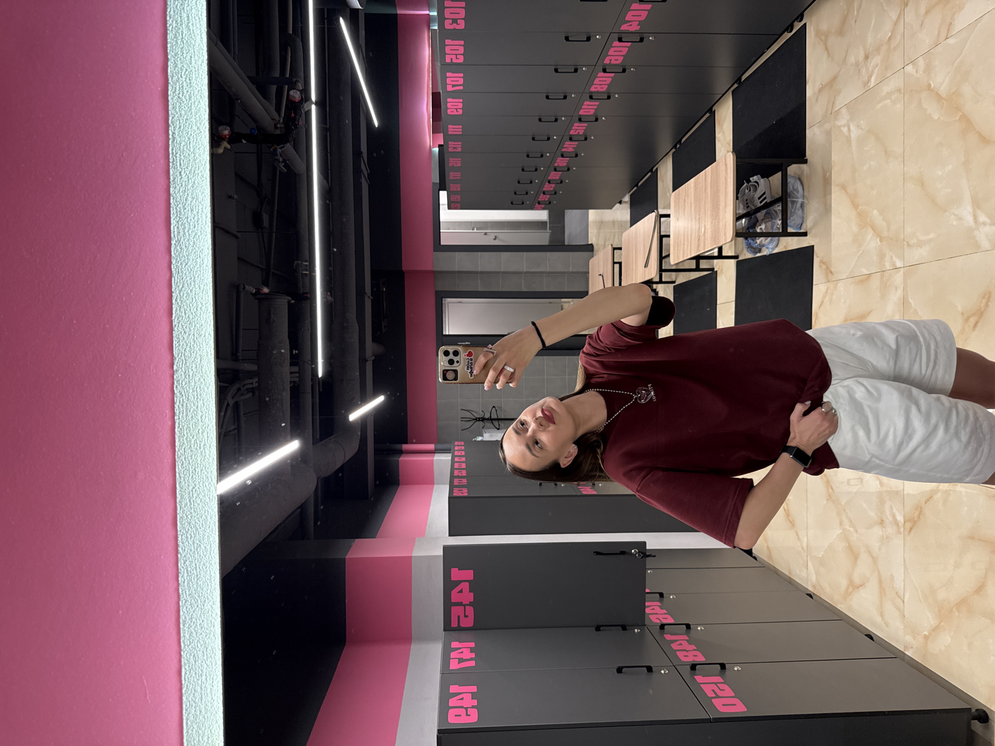 The image shows a young woman taking a mirror selfie in what appears to be a locker room or changing area. The room is decorated with bright pink and black accents, and the floor is tiled with a marble-like pattern. The woman is wearing a burgundy t-shirt and white shorts, and she has a heart-shaped pendant necklace. She is holding a smartphone in her right hand and wearing rings on her fingers. In the background, there are rows of dark gray lockers with pink lettering. Several wooden benches with black mats are placed between the lockers. The ceiling is black with fluorescent lights. There is no visible indication that this location is in Sochi, Russia.