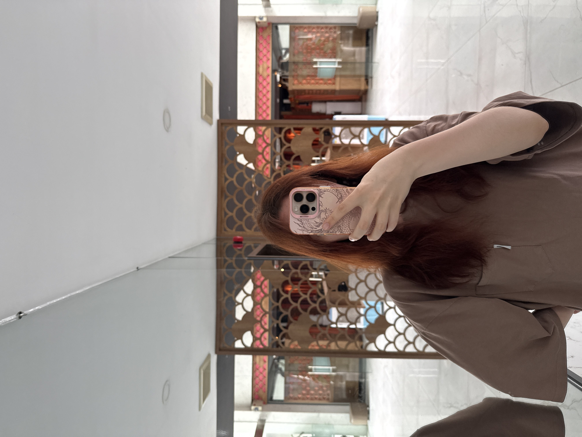 A person is standing in front of a mirror, holding a smartphone up to their face to take a picture or video of themselves. The phone has a pink case adorned with a dragon design. The person's face is obscured by the phone, and their auburn hair is visible around it. They are wearing a brown t-shirt.
The mirror reflects a doorway with decorative wooden latticework in a scalloped pattern. Through the doorway, a room with red accents and possibly stacked items can be seen. The surroundings appear to be an interior space, possibly a commercial establishment.
The location is identified as Bac Nha Trang Ward, Vietnam. The environment appears to be a modern building interior with marble flooring.