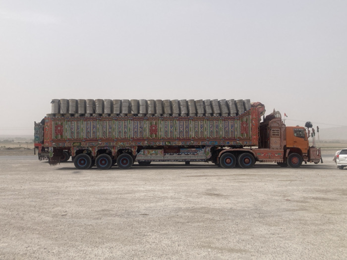 A large, brightly decorated truck is parked on a flat, dusty landscape under a hazy sky. The truck is loaded with many cylindrical objects wrapped in gray material. Ornate floral and geometric designs cover the truck bed and cab. A white car is parked near the truck's front. The overall scene suggests a transport vehicle in a dry, possibly arid environment.
