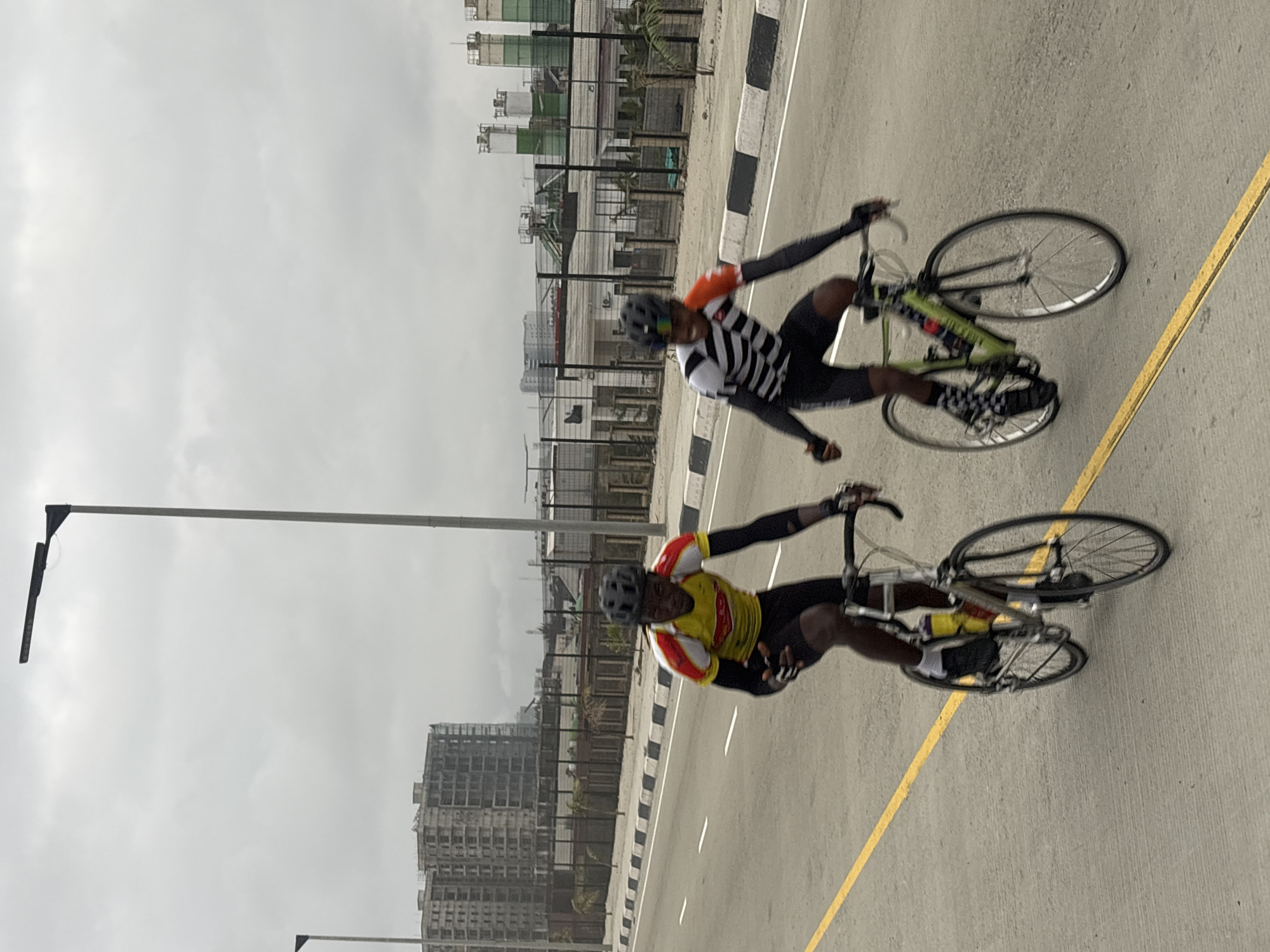 Two individuals are cycling on a paved road under a cloudy sky. The person in the foreground on the left wears a yellow and red cycling jersey with black shorts and a black helmet. They are actively pedaling and appear to be looking forward. To their right and slightly behind, a second cyclist in a black and white striped jersey with orange accents and a black helmet is also cycling, with their head turned towards the viewer. Both cyclists are on road bikes, with the front cyclist's bike being silver and the rear cyclist's bike being lime green.
The road is marked with a yellow line on the left and a white dashed line and solid white border on the right. In the background, there are various structures indicative of urban development or industrial activity. To the left, several tall, unfinished buildings are visible, some with scaffolding. Further in the background, to the right, there are industrial silos or storage tanks, some painted green. Streetlights are present along the road. The overall environment suggests an outdoor, potentially developing urban or industrial area in Alaguntan, Nigeria.