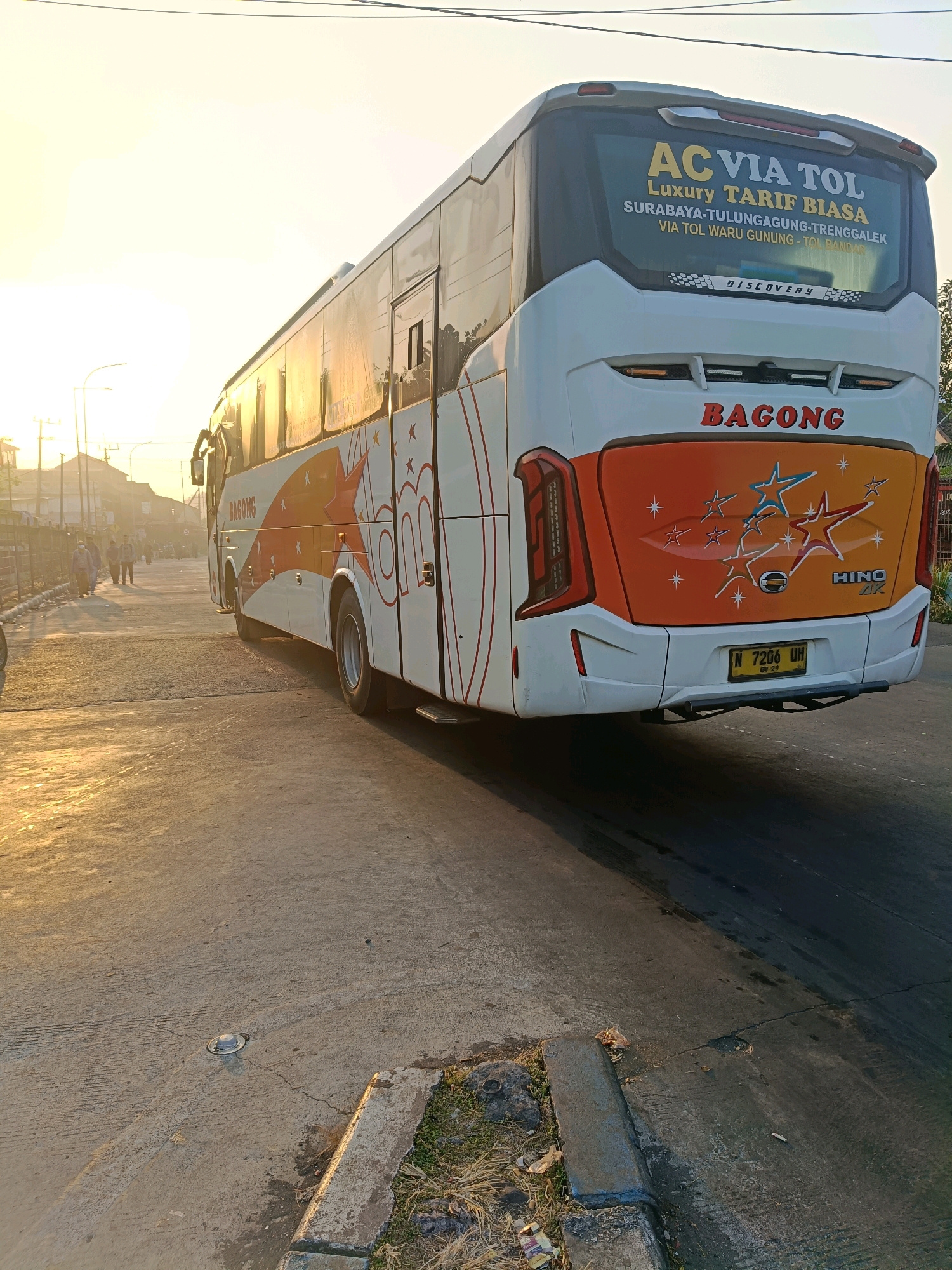 The image captures the rear of a white and orange "BAGONG" intercity bus parked on a road in Kedungrejo, Indonesia. The bus displays "AC VIA TOL TARIF BIASA" and route information "SURABAYA-TULUNGAGUNG-TENGGALEK VIA TOL WARU GUNUNG - TOL BANDAR" on its rear window. A HINO AR emblem is visible on the orange section of the bus, along with a star graphic. The bus has license plate number "N 7206 UH". The scene is illuminated by the warm, low light of either sunrise or sunset, casting long shadows. In the background, several individuals are walking on the side of the road, with buildings and utility poles visible. The road surface is asphalt with visible cracks and some debris, including a discarded cigarette butt and some plastic waste in the foreground. A metal grate is present on the road surface.