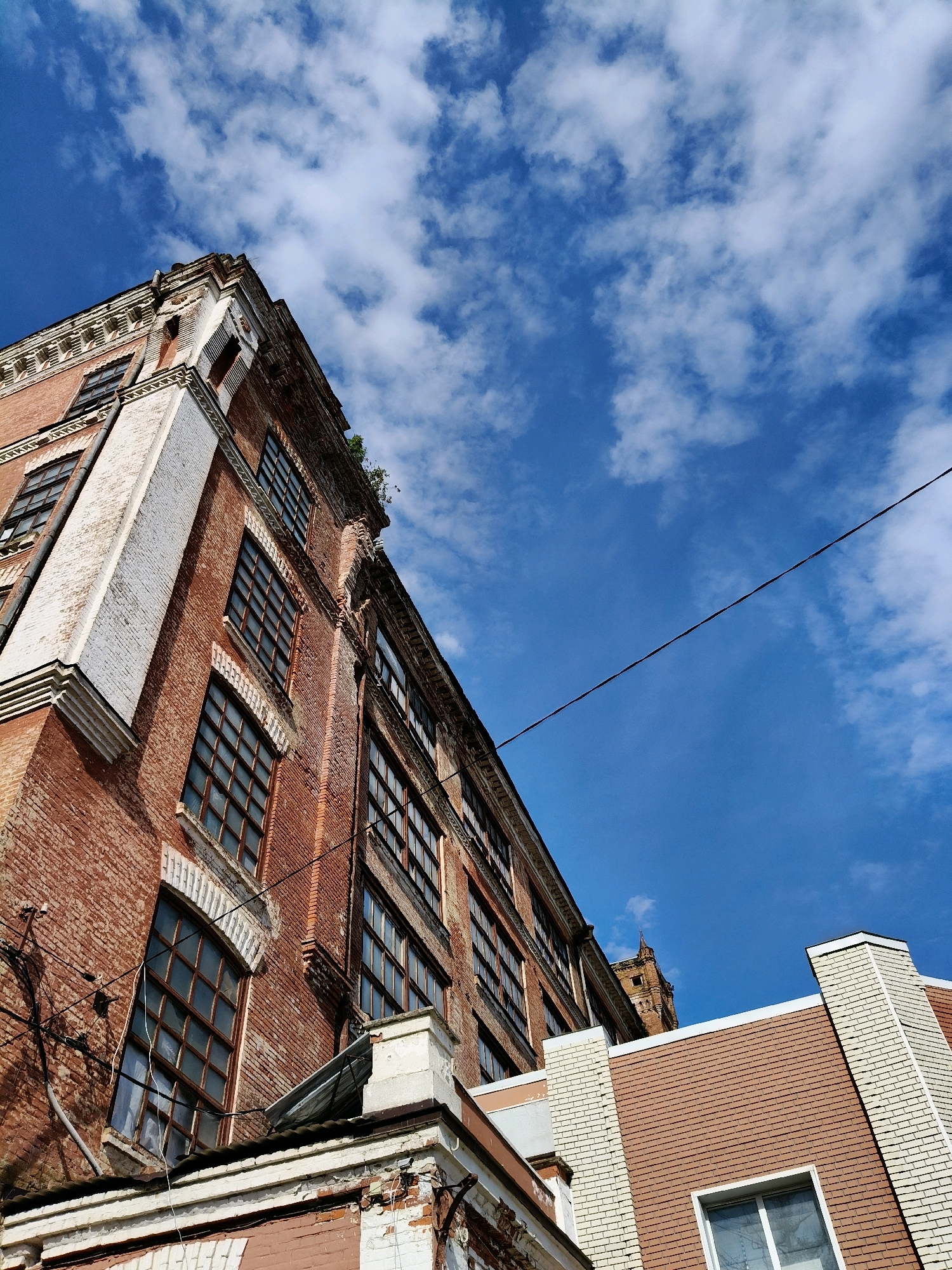 The image shows an upward view of brick buildings against a blue sky with scattered clouds. The main building in the foreground is made of red brick and features numerous windows with dark frames. Its architecture suggests it is an older structure, possibly industrial or historical, with white decorative elements along its cornices and corners. A dark wire stretches diagonally across the sky. In the background, another building with a distinct tower and a more modern brick facade is partially visible. The overall impression is that of an urban industrial or historic district under a bright sky. This scene is located in Naro-Fominsk, Russia.