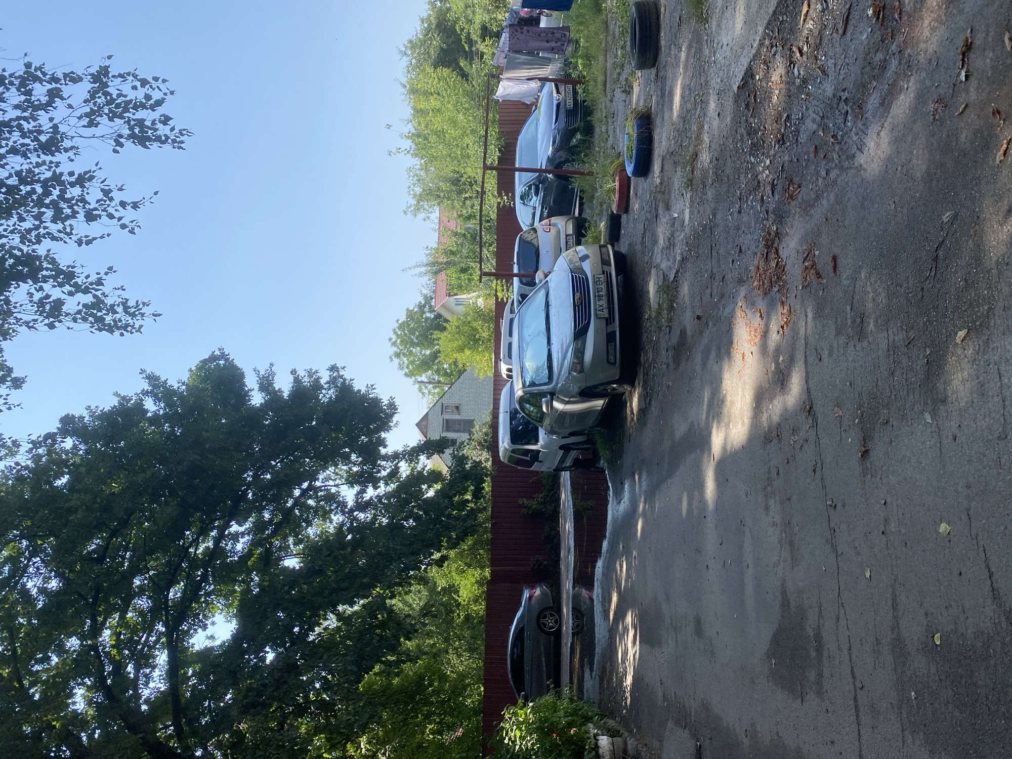 The image depicts an outdoor parking area with several cars. In the foreground, a cracked asphalt surface is visible, with puddles of water and scattered leaves. A dark gray sedan is parked on the left, with its reflection visible in a large puddle. To the right of this sedan, a white minivan and a silver sedan are parked. Further into the scene, a dark gray SUV is parked. In the background, a red wooden fence runs horizontally across the image. Beyond the fence, houses and trees are visible. A clothesline with laundry hanging on it is also present. The sky is clear and blue. The scene appears to be in a residential area of Poltava, Ukraine, likely a backyard or communal parking space.