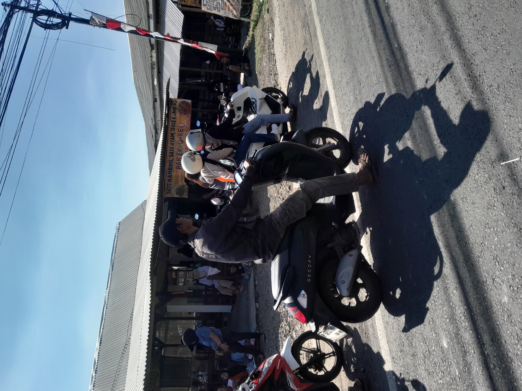 In Gentengwetan, Indonesia, on a sunny day, people are seen on a roadside. A man wearing a dark cap and jacket rides a dark scooter, his shadow cast on the asphalt road. To his left, another scooter, red and white, is parked. Further back, under a corrugated metal roof structure, several people are seated. A banner advertising "SATE KAMBING, SATE AYAM, GULE KAMBING PAK EK-O-KE" is visible. Another white scooter carries two individuals wearing helmets. The environment is outdoors with a clear blue sky above.