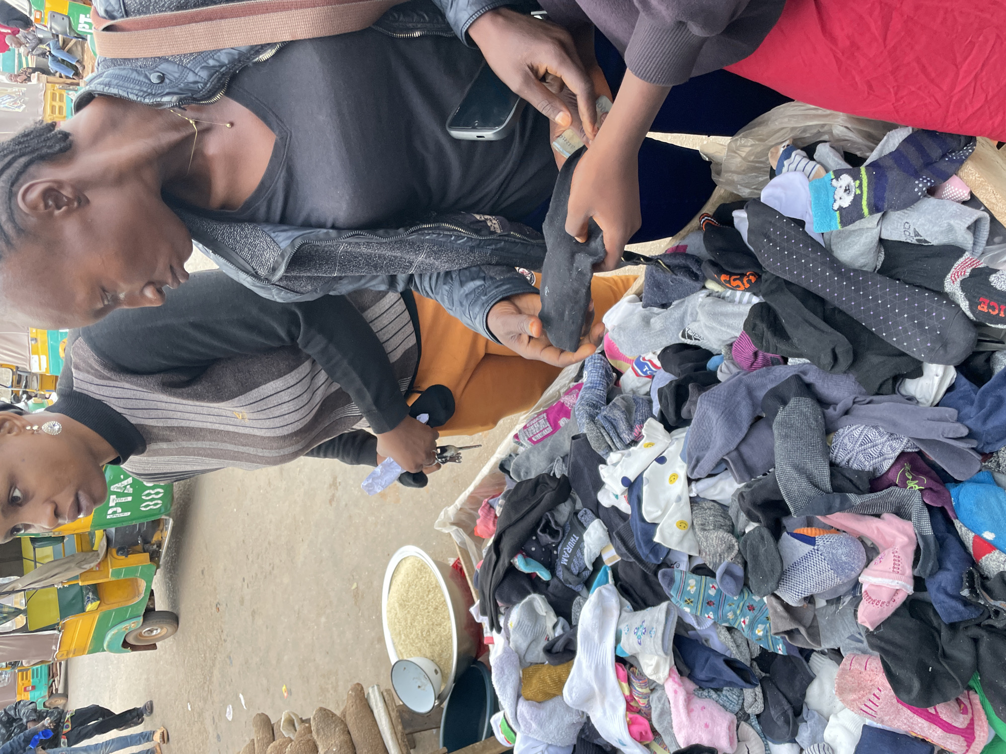 This image captures a vibrant street market scene in Gura Top, Nigeria. In the foreground, three individuals are positioned around a merchandise display. On the left, a woman wearing a black and brown striped turtleneck sweater and orange trousers holds keys and a small white object in her hands. In the center, a woman dressed in a black V-neck shirt, a black textured jacket, and dark blue trousers, holds a black sock in her left hand. Her right hand, also holding a black smartphone, is extended towards the hand of a third, partially visible individual on the far right, who is holding banknotes. The third individual wears a dark purple sleeve and a red garment.
The central focus of the merchandise is a substantial pile of assorted socks of various colors, patterns (including smiley faces, stripes, and dots), and sizes, contained within a large box. To the left of the socks, a large metal bowl filled with light-colored grains, resembling rice, is visible, alongside a smaller white enamel bowl with a blue rim. Behind these items, a stack of elongated, brown, rough-textured produce is arranged. In the background, a green and yellow three-wheeled auto-rickshaw (Keke Napep) with the partial text "PTA-" and "88" visible on its side, is parked on what appears to be a paved or dirt road. Other people and structures are discernible in the distance, indicating an active outdoor market environment.