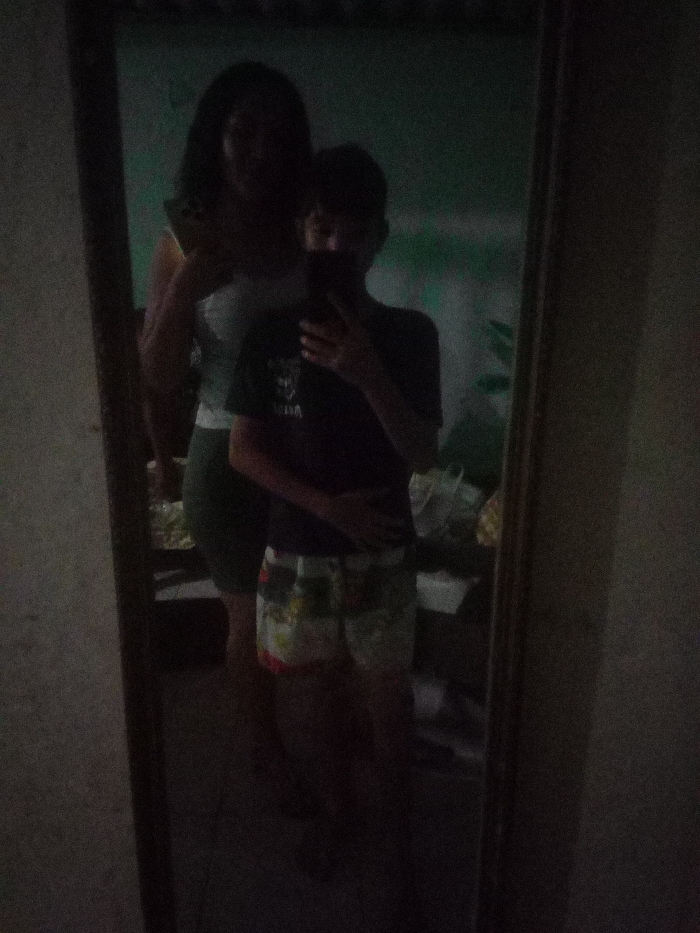 A dimly lit selfie captured in a mirror reflects two figures. A taller woman stands behind a person holding the camera, obscuring their face. The woman wears a light-colored top and a darker skirt. The selfie-taker is dressed in a dark t-shirt and patterned shorts. The background appears to be a room, with indistinct shapes and light patches. The photo has poor lighting and a grainy quality.
