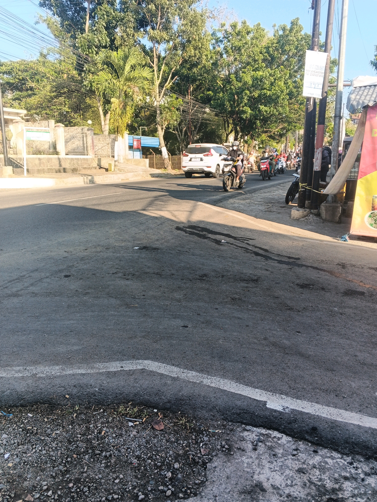 The image depicts a street scene in Semarang, Indonesia, likely during daylight hours due to the shadows cast by the trees and buildings. The foreground is dominated by a grey asphalt road with a white lane marking running diagonally from the bottom left to the upper right. The road surface shows signs of wear, with some dark patches and debris scattered on it, including small stones and what appear to be cigarette butts near the curb.
To the left of the road, there is a raised concrete sidewalk and a low stone wall with a fence. Behind the wall, a building with a white facade and a blue roof is partially visible, along with lush green trees and foliage. Overhead, a tangle of power lines crisscrosses the sky.
The middle ground of the image shows a moderate flow of traffic. A white SUV is visible on the road, and several motorcycles are also present, some with riders. One motorcycle, closer to the viewer, has a rider wearing a dark helmet and a light-colored shirt.
To the right of the road, there is a tall utility pole with a white sign attached to it, featuring some text that is difficult to read. Beyond the pole, part of a building with a yellow and red facade is visible, possibly a storefront or market stall. More trees and buildings can be seen in the background, under a clear blue sky. The overall impression is of a typical urban street in a tropical or subtropical climate, characterized by a mix of infrastructure, vegetation, and vehicular activity.