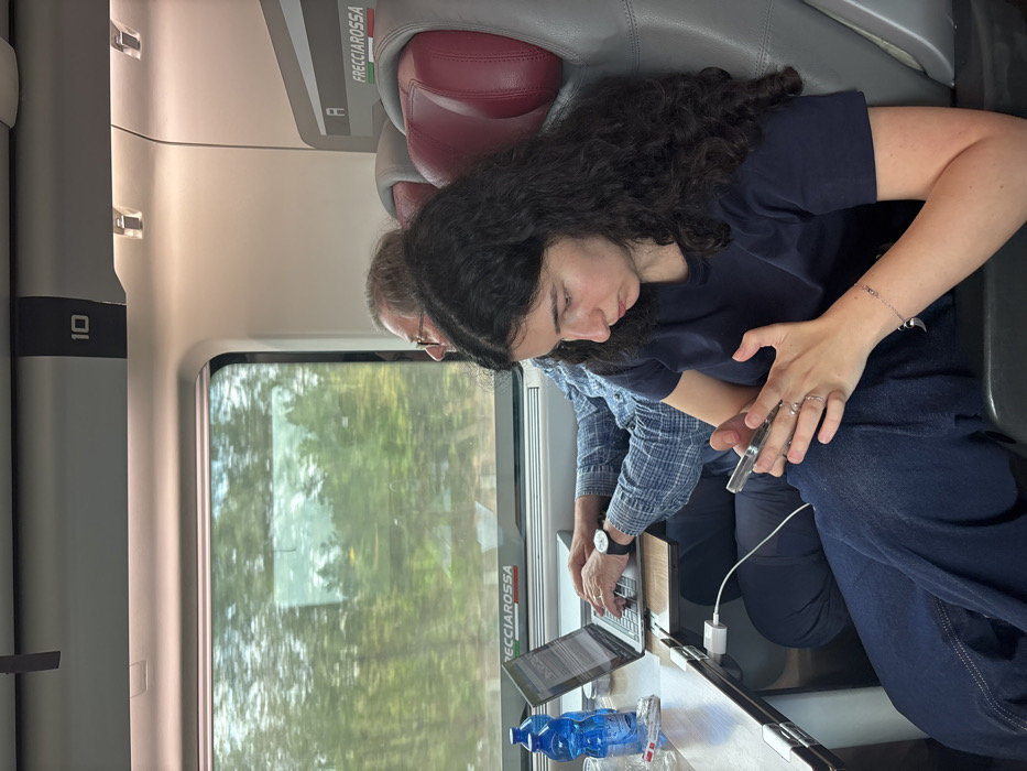 The scene depicts the interior of a Frecciarossa high-speed train carriage, likely traveling through or near Castelfranco Emilia, Italy. Two individuals are seated. In the foreground, a young woman with long, dark, curly hair, wearing a dark blue t-shirt and dark blue pants, is engrossed in a silver smartphone held in both hands. She has rings on her left hand and a silver bracelet on her right wrist. Behind her and to the left, an older man with short, gray hair and eyeglasses, dressed in a blue plaid shirt, is seated at a wooden fold-out table next to a window. He is actively typing on an open silver laptop. A blue plastic water bottle, a clear glass, and a small metallic container rest on the table. A white charging adapter with a cable is plugged into a power outlet below the table. The man wears a watch on his left wrist. Through the train window, a blurred exterior shows green trees, consistent with a train in motion through a natural landscape. The train interior features grey seats with red headrests and an overhead panel marked with the number "10".