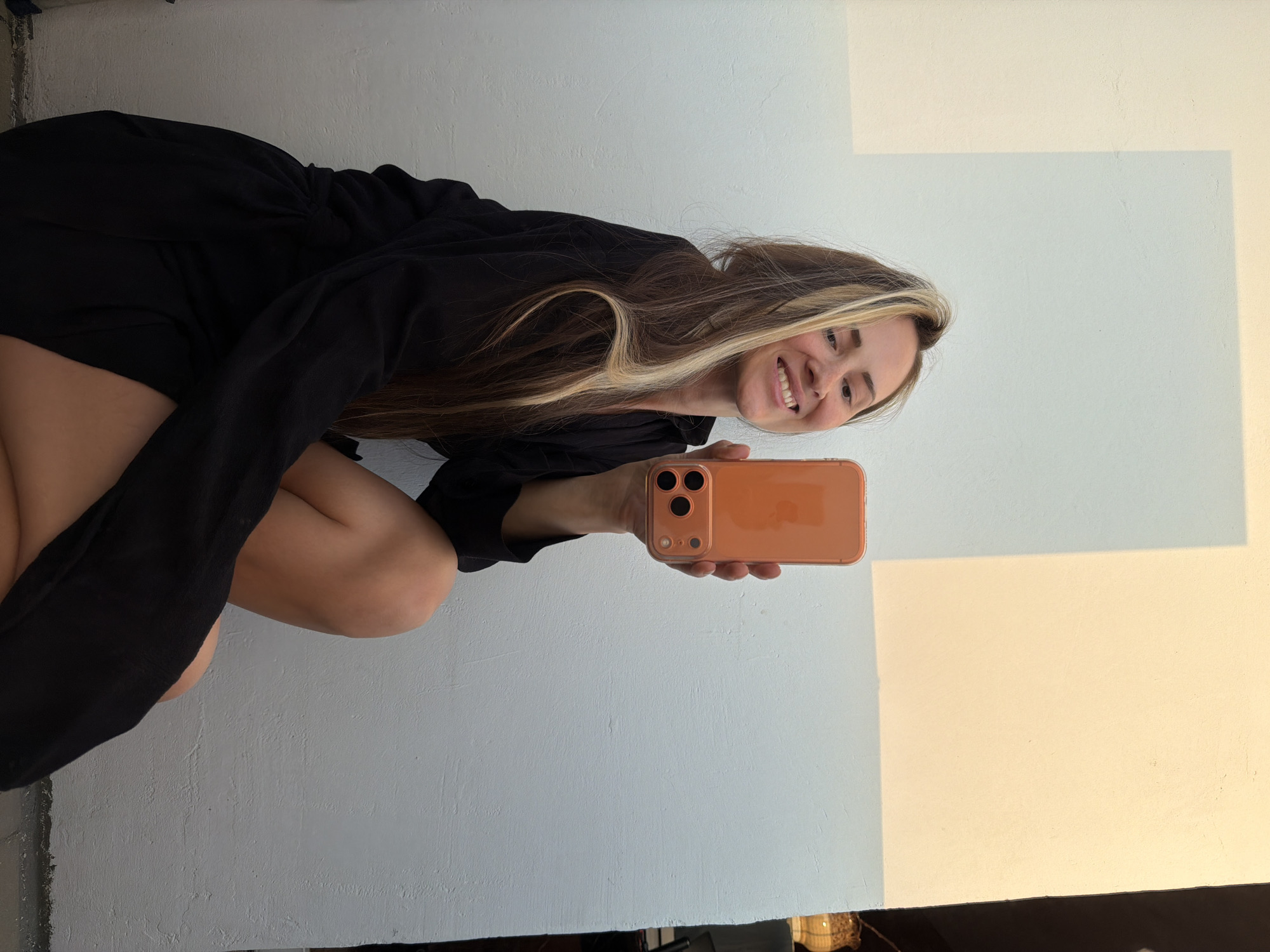A woman with long brown hair with blonde streaks, wearing a black garment, is squatting and holding a peach-colored iPhone in front of her. She is smiling at the camera. The background is a two-toned white wall. The location information is not determinable from the image.