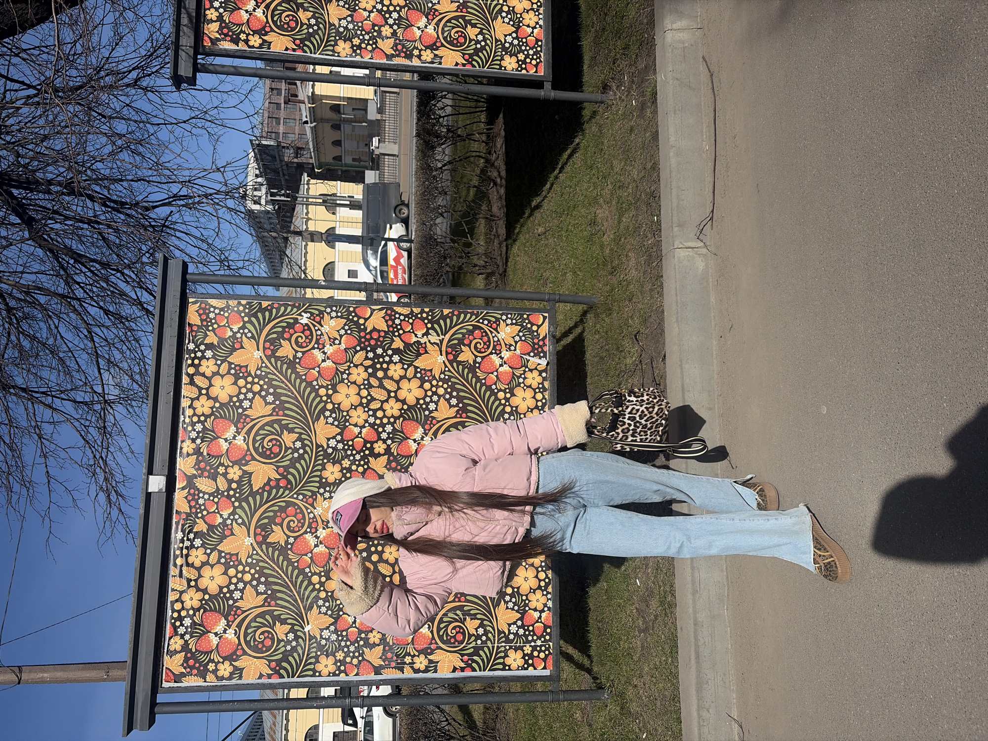 A young woman lies on a bench displaying a patterned advertisement in Saint Petersburg, Russia. The bench features a floral design with red strawberries, yellow flowers, and green leaves against a black background, framed by a dark metal structure. The woman, wearing a pink jacket, light blue jeans, and a baseball cap, poses for the camera. A leopard-print handbag sits on the ground near her feet. The surrounding environment includes bare trees, green grass, a concrete sidewalk, and buildings in the background. A vehicle is visible in the distance, adding to the urban context.