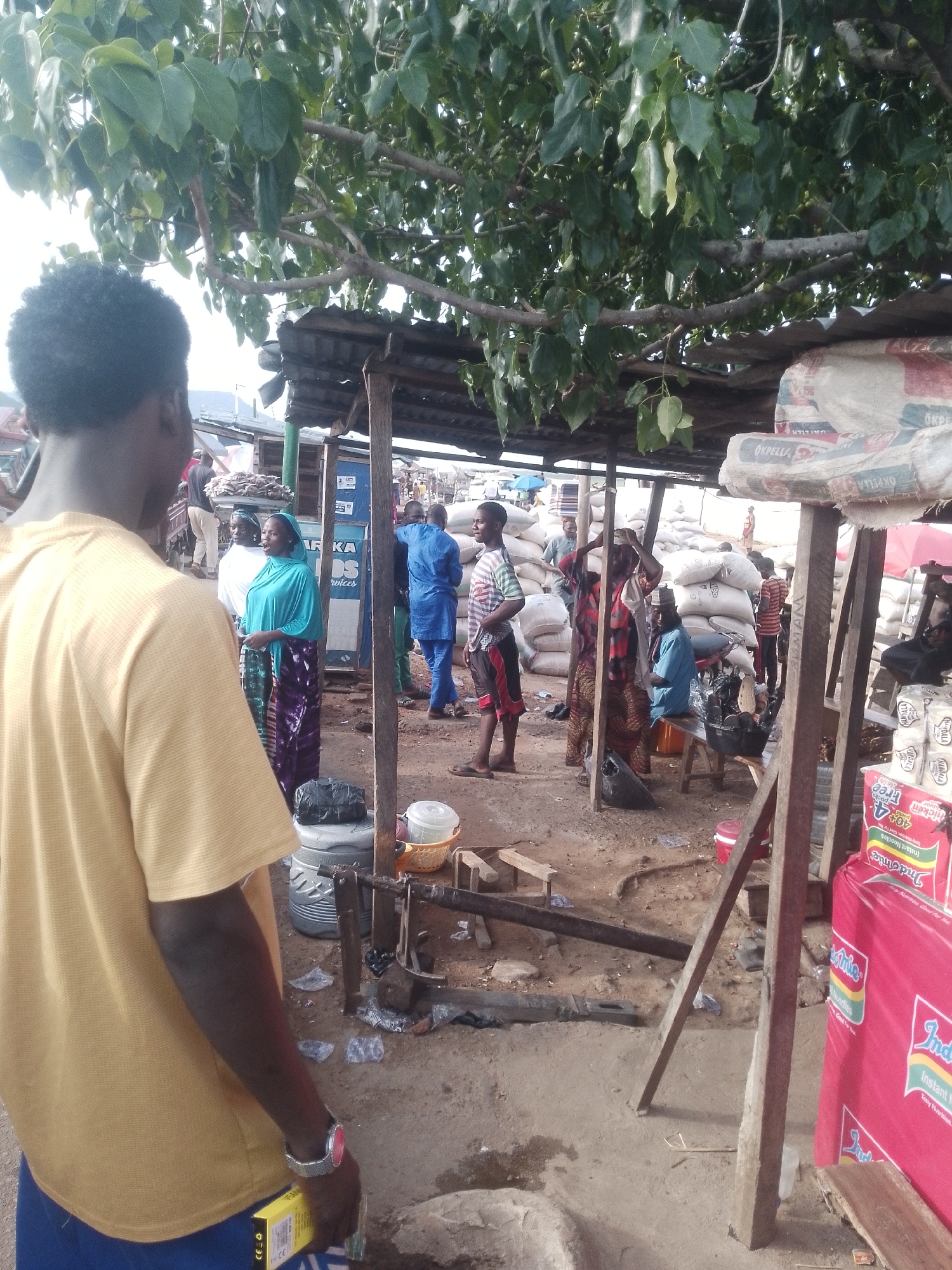 The image shows a bustling outdoor market scene. In the foreground, a man with short black hair and a yellow shirt stands with his back to the camera, obscuring a portion of the view. He wears a wristwatch on his left wrist and holds a small yellow box.
The market area is sheltered by a simple corrugated metal roof supported by wooden beams. Several individuals are visible beneath the shelter: a group of women in traditional clothing stand near a sign. A man in a blue outfit interacts with others. People are milling around, and some appear to be carrying sacks of goods.
In the background, there are piles of large white sacks labeled "OKPELLA". There's also a red display stand for "Indomie" instant noodles. The ground appears to be dusty or sandy. A set of scales and a variety of containers and supplies are scattered on the ground. A tree with broad green leaves overhangs the scene, providing some shade.