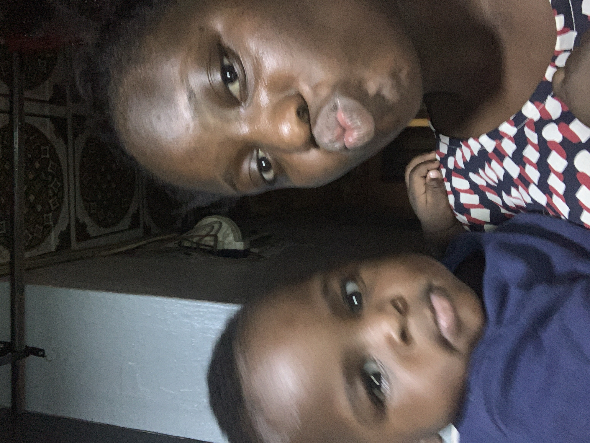 In a close-up, dimly lit selfie, a young Black woman and a baby are visible. The woman, on the right, is making a kissing face, her lips puckered. She has dark skin and dark hair, with some facial highlights suggesting she might be wearing makeup. Her patterned shirt, featuring a navy blue background with white and red oval shapes, is partially visible. To the left, the baby is looking towards the camera with a neutral expression. The baby has dark skin and is wearing a dark blue shirt. The background is blurred and dark, with hints of a textured ceiling above and a light-colored wall to the left. The overall lighting is low, with harsh light creating strong reflections on the woman's face. The scene appears to be indoors, with no identifiable landmarks or specific locations within the city of Unknown, Unknown.