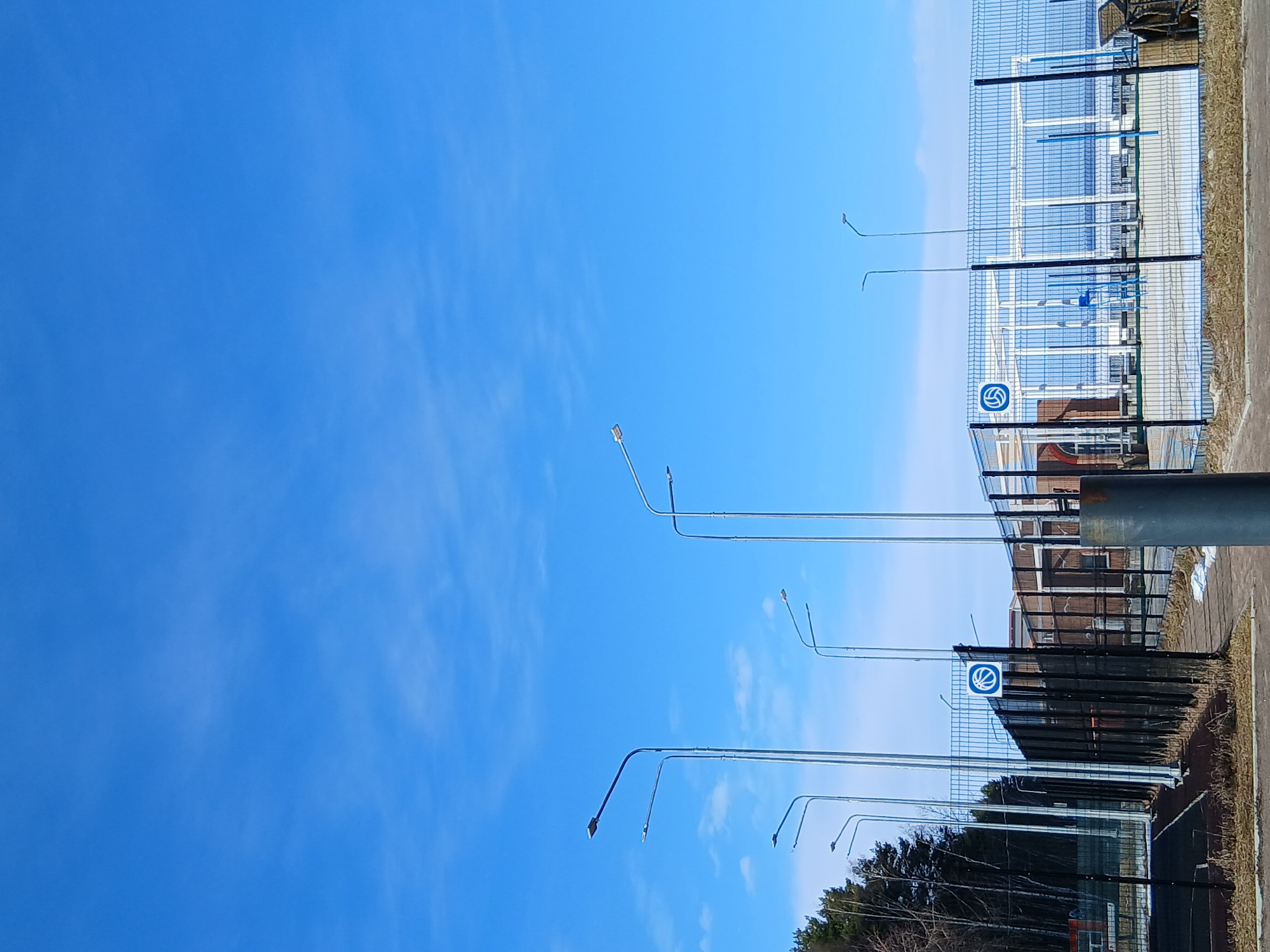 The image shows a basketball court in an unknown location. The sky is mostly blue. There are light poles extending upward along the edges of the court. A fence with a basketball logo is visible. The ground around the court is a mix of pavement and dirt.