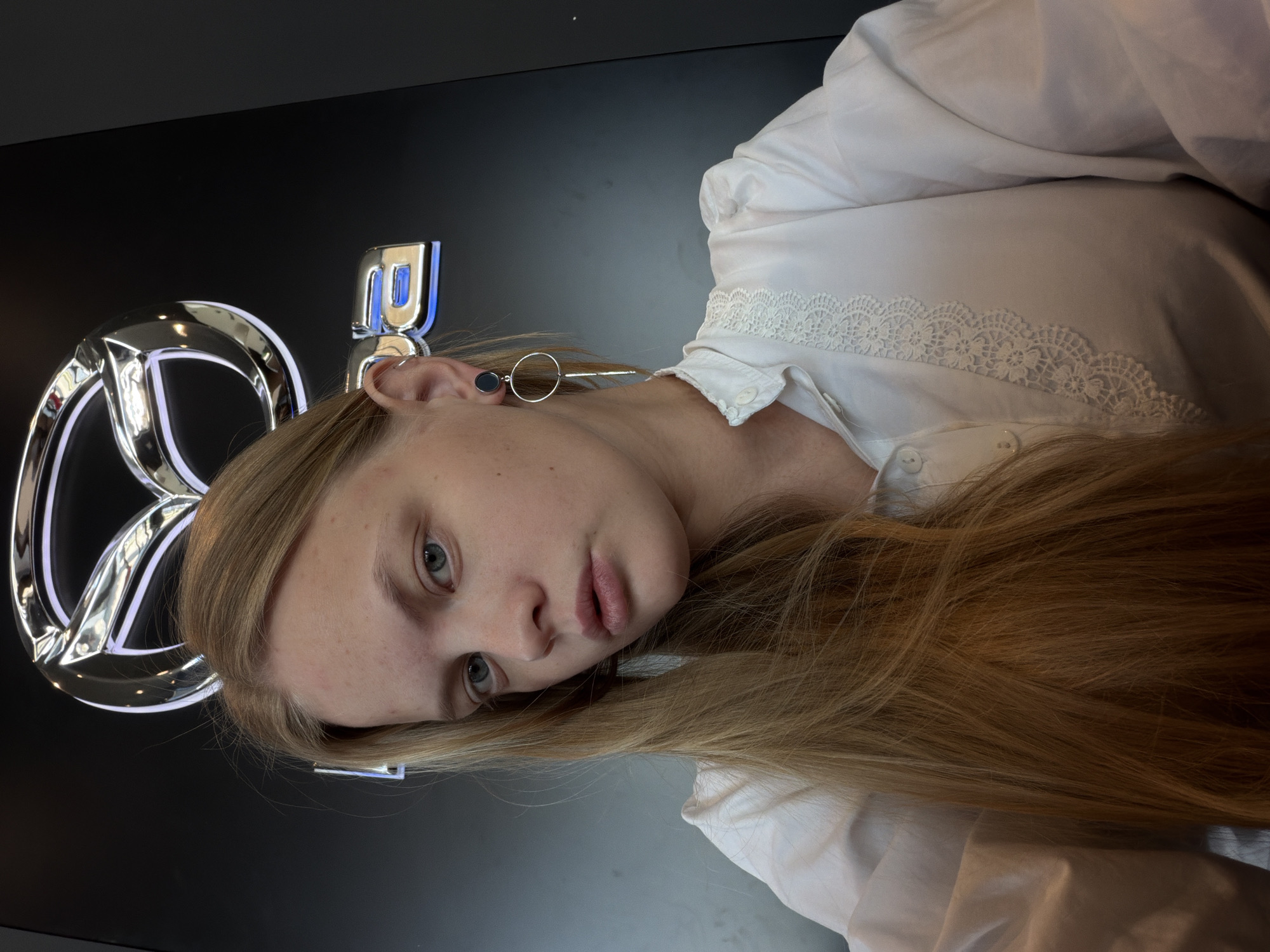 A medium close-up, rotated portrait photo captures a young individual, likely female, with long, straight, blonde hair and blue-grey eyes, gazing directly at the camera with a neutral expression. She wears a white long-sleeved shirt adorned with intricate white lace detailing along the placket. A distinctive earring is visible in her left ear, featuring a small black stud connected to a larger, extending silver circular hoop.
The background is a dark, possibly black, surface, prominently featuring a large, illuminated chrome logo. The visible portion clearly identifies the stylized "M" within an oval, characteristic of the Mazda car brand, outlined with blue or white light. To the right of the main logo, a partial view of another chrome, blue-lit letter or symbol is visible, possibly part of the brand name "MAZDA".
The setting appears to be indoors, likely a car showroom, dealership, or an event associated with the Mazda brand, located in Izhevsk, Russia, as suggested by the context. The lighting is artificial, highlighting the metallic sheen of the logo and the subject's features. The activity is a casual pose for a photograph, possibly a selfie.