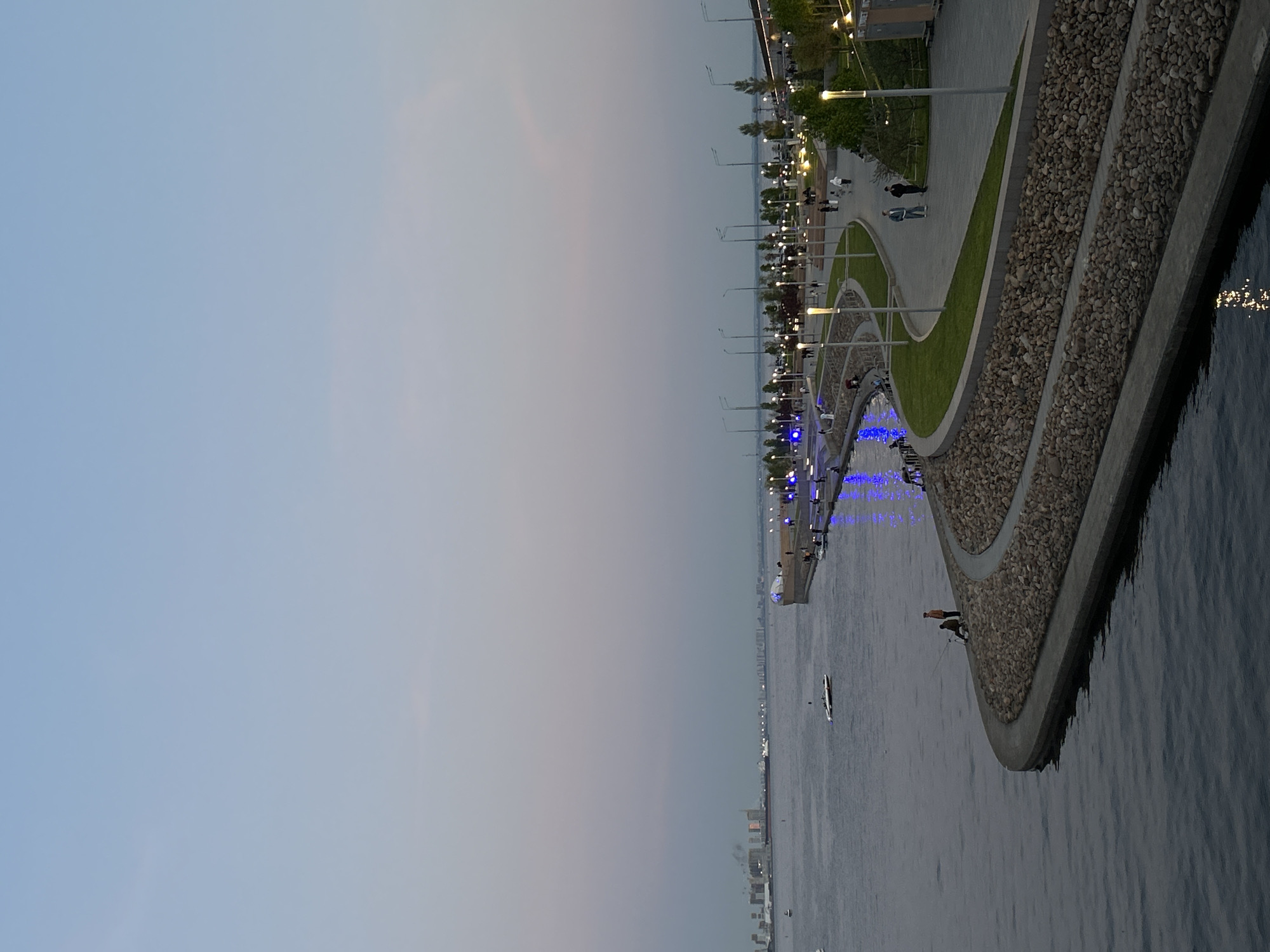 Here is a description of the image:
The image shows a long concrete embankment along a river or large body of water, which is likely the Neva River in Saint Petersburg, Russia. The embankment features a walkway with lampposts and green spaces, including strips of grass. There is a gravel border next to the water. People are visible walking along the walkway. Blue lights are visible along the embankment near the water. A boat is on the water. The sky is overcast, with varying shades of light blue and grey. Buildings are visible in the distance along the water. Two people are fishing on the embankment.