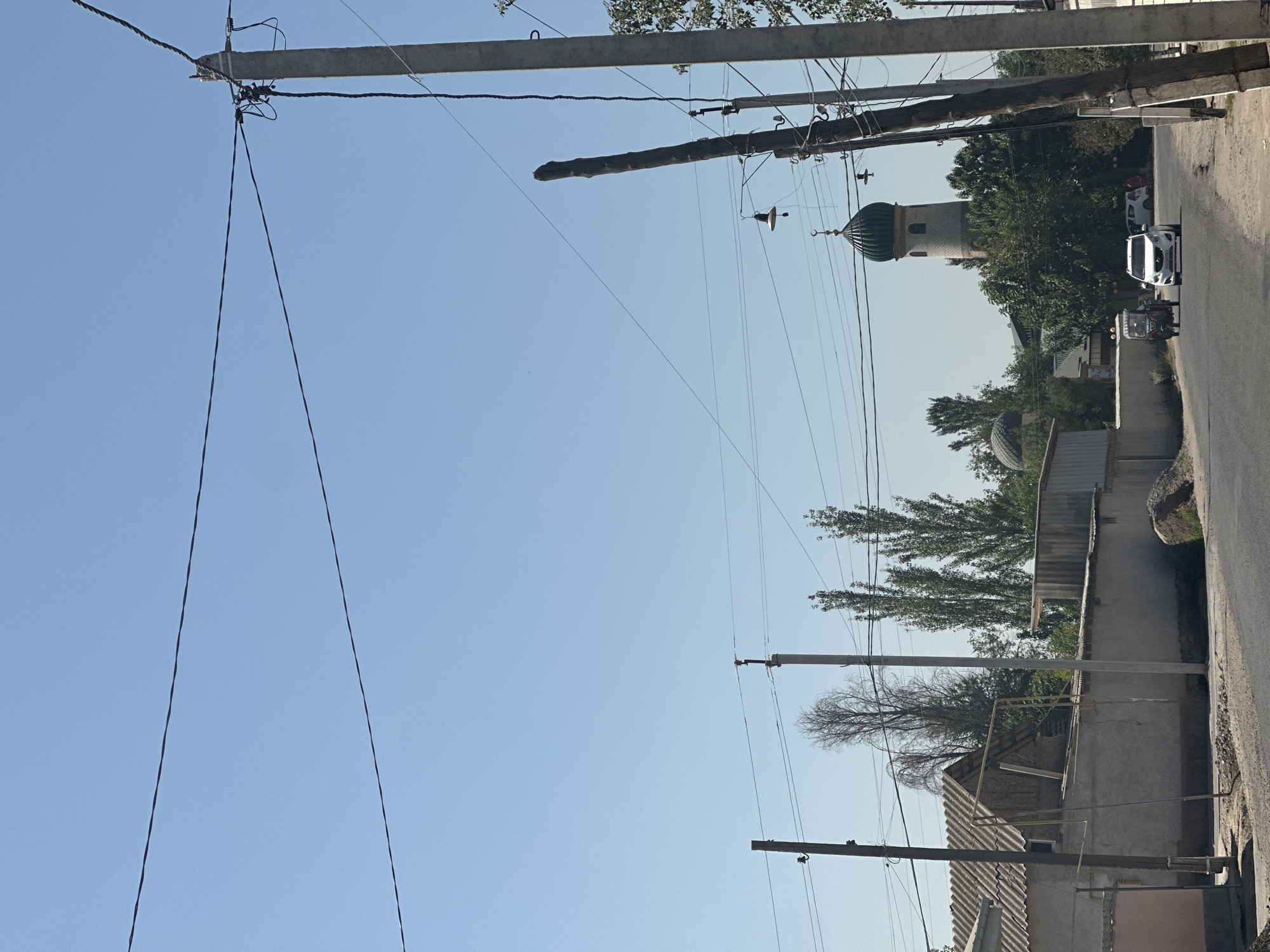 The image depicts a street scene in Yallama, Uzbekistan, under a clear blue sky. On the left, a low-rise building with a corrugated roof is visible, bordered by a light-colored wall. In the distance, a prominent mosque with a green dome and a tall minaret stands out against the trees. A white SUV and a three-wheeled auto-rickshaw are driving on the asphalt road. Utility poles and wires crisscross the upper portion of the image. The overall environment appears to be a rural or semi-urban area with a focus on local architecture and infrastructure.