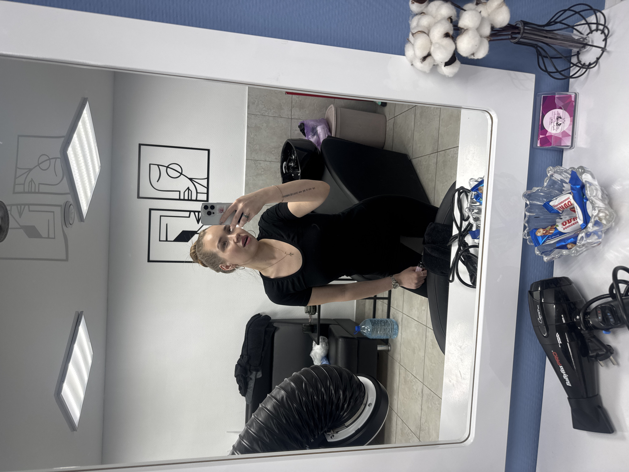 A woman is taking a selfie in a mirror in what appears to be a salon or beauty parlor. The location is likely Chaikovsky, Russia, due to the presence of Russian text on the candy packaging. The mirror reflects a room with salon equipment, including chairs, a sink, and ventilation. Artwork is on the wall behind the woman. In the foreground, on the right, are decorative items such as cotton branches, a candy dish with Russian-branded chocolate, a salon business card, and a hair dryer. The woman is dressed in black and holding a smartphone.