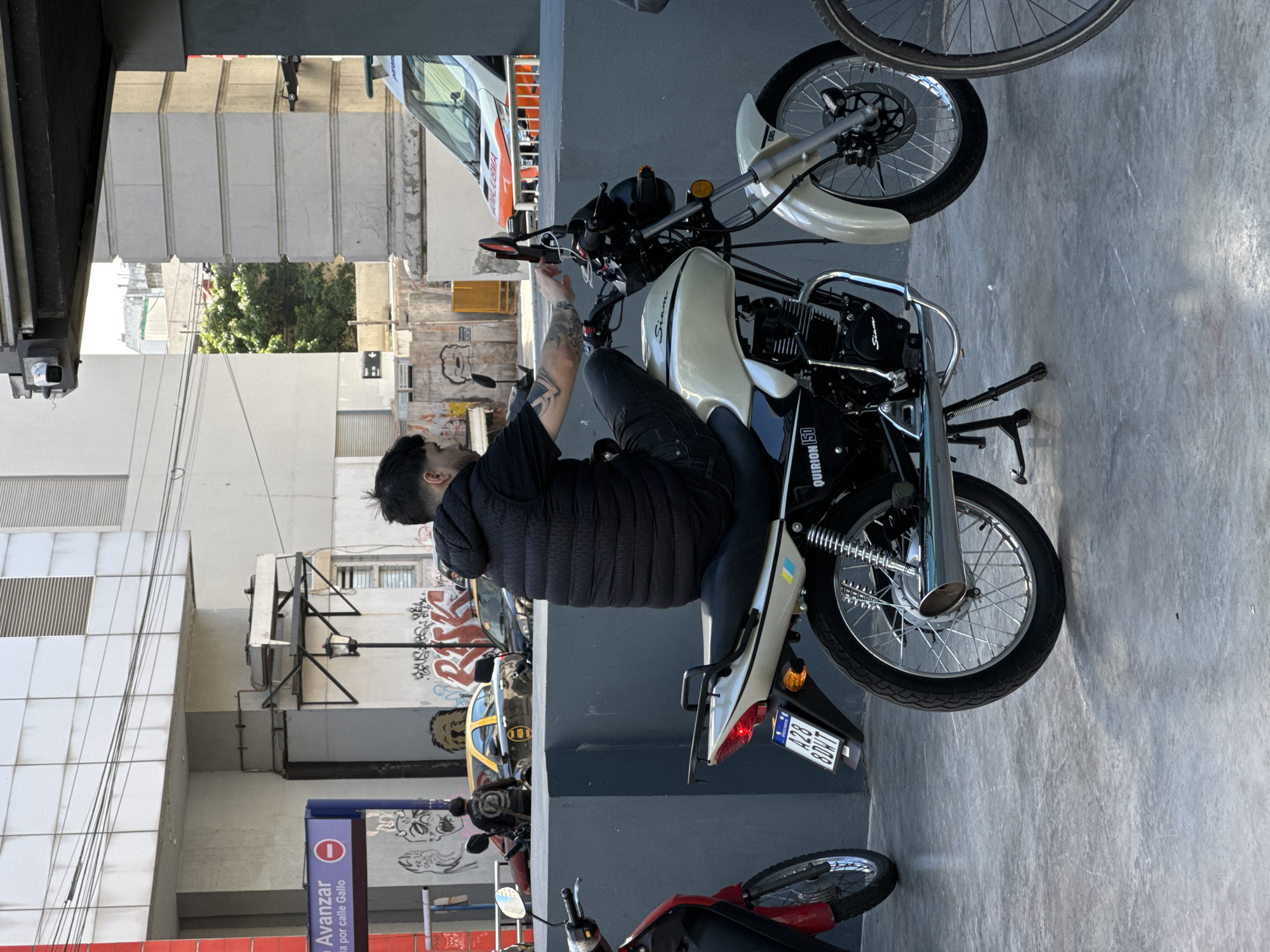 The image depicts a man on a white and black motorcycle in a city environment. The man is wearing a black jacket and dark pants. He is sitting on the motorcycle and appears to be adjusting something on the handlebars. The motorcycle is parked on a concrete surface.
In the background, there is a building with graffiti on its walls, a blue sign that reads "Avanzar," and several other parked vehicles, including a taxi. Based on the "Avanzar" sign and the context, the location is likely in Buenos Aires, Argentina.
