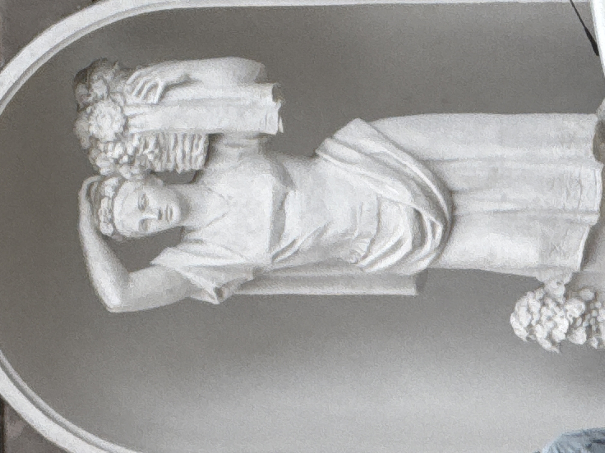 The image depicts a white stone statue of a woman carrying a basket of flowers on her head. The statue is positioned within an arched alcove, with a plain gray background. The woman is wearing a flowing robe, and her left arm is raised to support the basket. To the lower left of the main statue, a smaller, less detailed sculpture of a floral arrangement is visible. A portion of a blue garment, possibly clothing on a person, is also visible at the bottom left edge of the frame, partially obscured by the smaller statue. The overall scene is monochromatic and appears to be an outdoor or semi-outdoor architectural feature. The statue's style suggests a classical or neoclassical influence. It is not possible to determine the specific location in Moscow, Russia from the visual information provided.