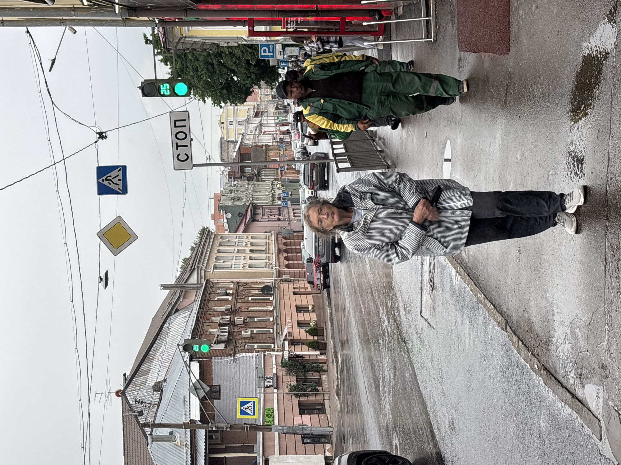 The image depicts a street scene in Nizhny Novgorod, Russia, on a cloudy and wet day. A woman wearing a striped jacket and dark pants is walking on a wet sidewalk in the foreground, facing the camera. Behind her, two men in work uniforms, one in green and yellow and the other in a darker uniform, are walking in the same direction. Several cars are visible on the wet street, and buildings line the right side of the road. Traffic lights and street signs, including one in Russian that reads "СТОП" (STOP), are present overhead. The overall atmosphere is urban and somewhat dreary due to the overcast weather and wet conditions.