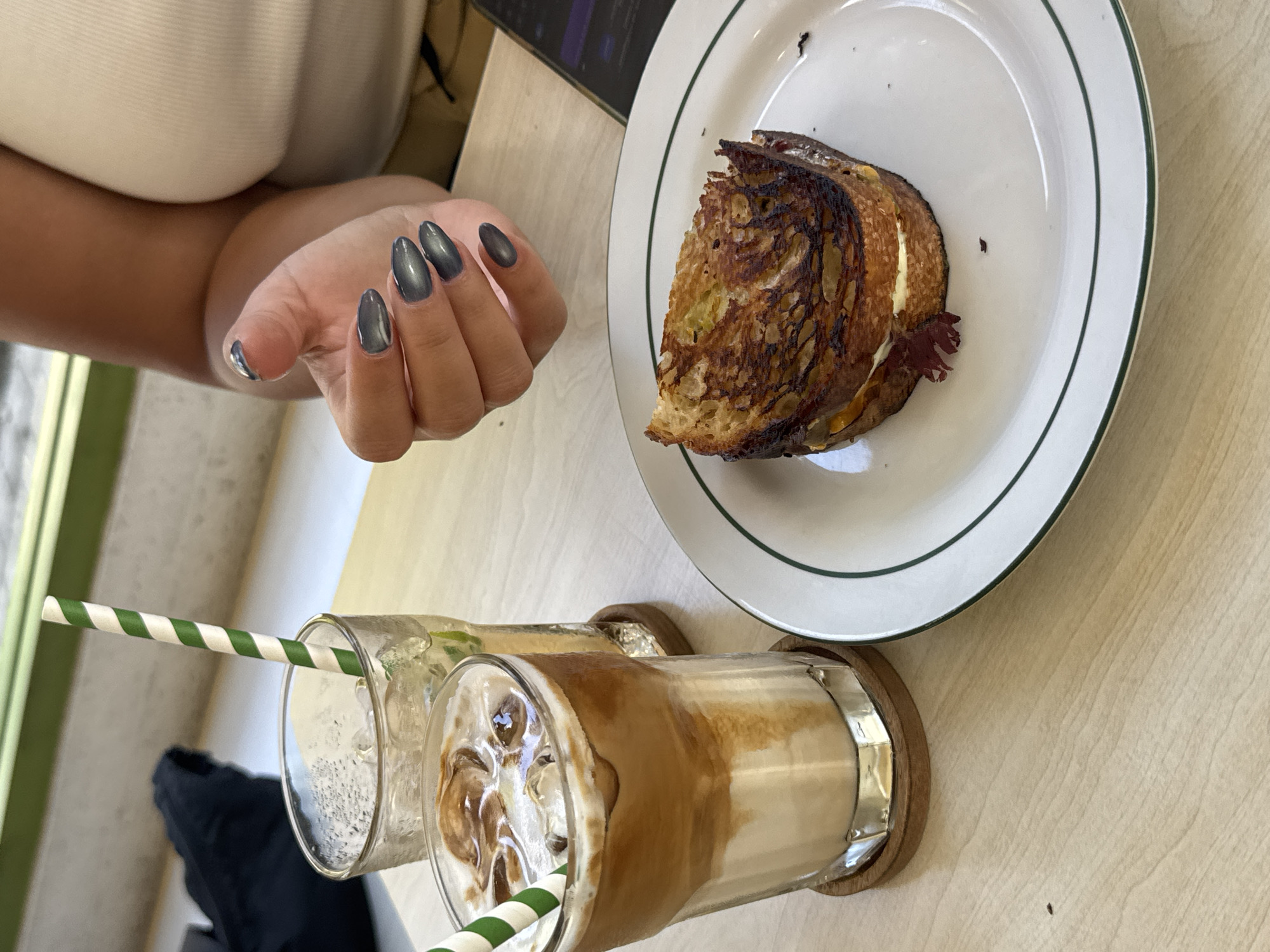 The image depicts a meal setting, likely in North Kuta, Indonesia. A person's hand with grey almond-shaped nails is visible to the left. In the upper right, there is a piece of toasted sandwich on a white plate with a green rim. Two glasses containing beverages, one clear and one with coffee layered over milk and ice, sit on wooden coasters near the bottom of the frame. Both have green and white striped straws. The table is light-colored and appears to be made of wood or a wood-like material. The background is indistinct, but a glimpse of a green vertical surface can be seen on the left.