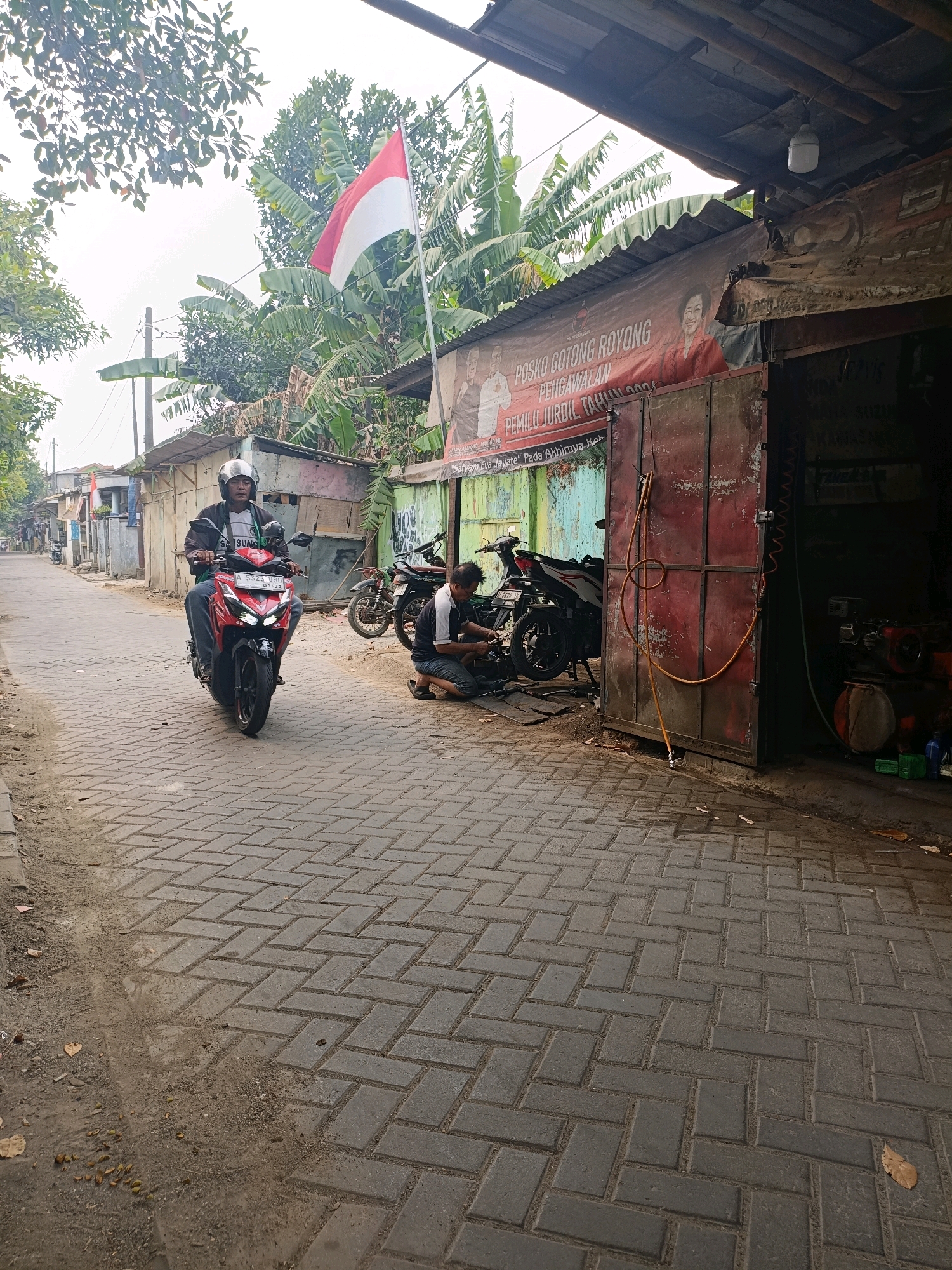 In Tangerang, Indonesia, a man rides a red motorcycle down a brick-paved street, wearing a helmet and a dark jacket. The street is lined with small buildings. To the right, a workshop with a large red metal door is open, revealing a mechanic working on a motorcycle. A red banner with Indonesian text and images of people hangs above the workshop. A red and white Indonesian flag flies from a pole in the background, with banana trees and other foliage visible. The sky is overcast.