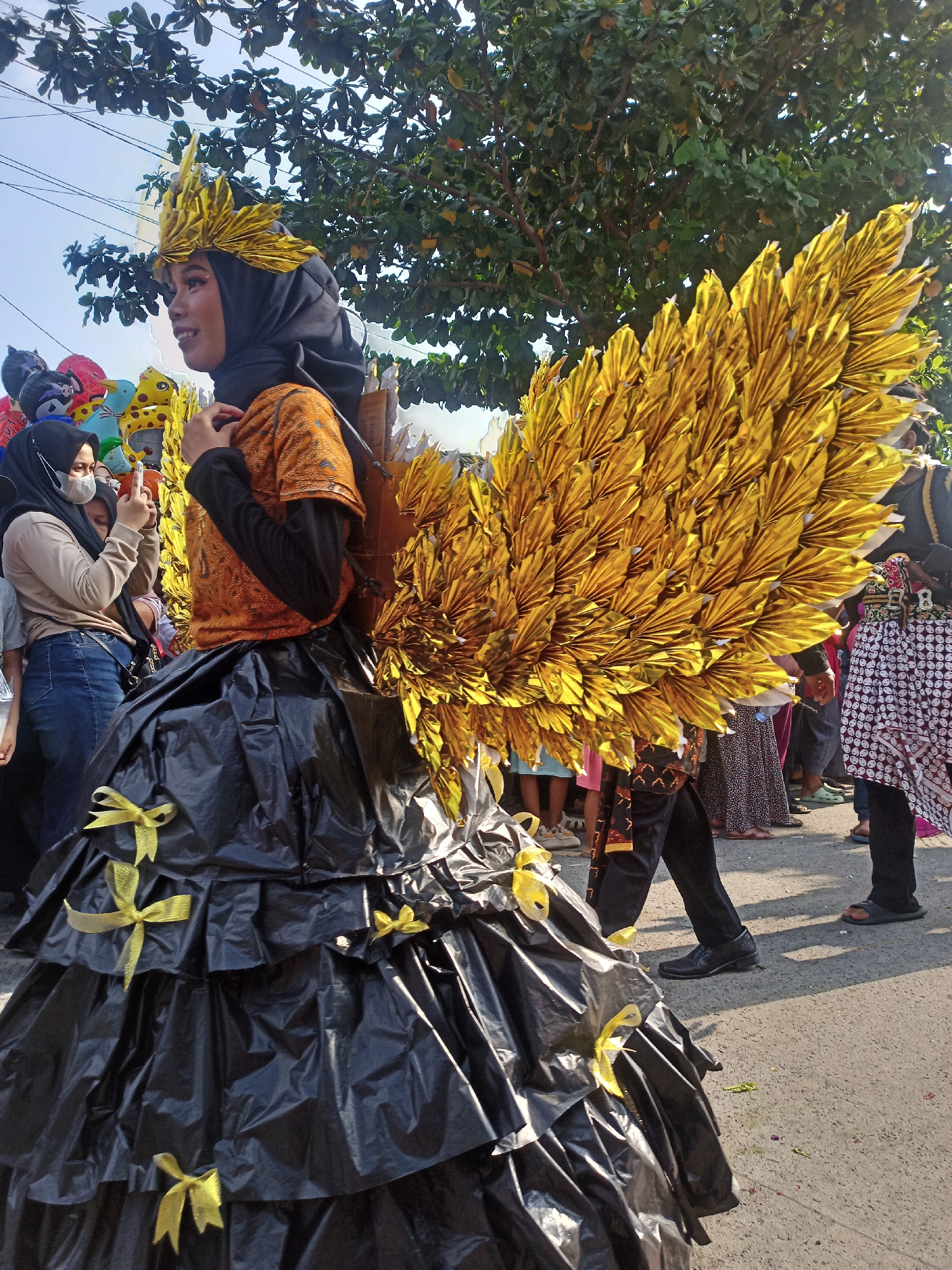 In Saren, Indonesia, a young woman participates in a parade. She is wearing a black hijab and an orange top with gold embellishments. Her elaborate costume features a voluminous black skirt made of gathered material, adorned with yellow bows, and a pair of large, golden, feather-like wings. She is also wearing a golden headpiece that resembles a crown of feathers. In the background, other parade participants and onlookers are visible, along with trees and balloons. The event is taking place outdoors on a paved street.