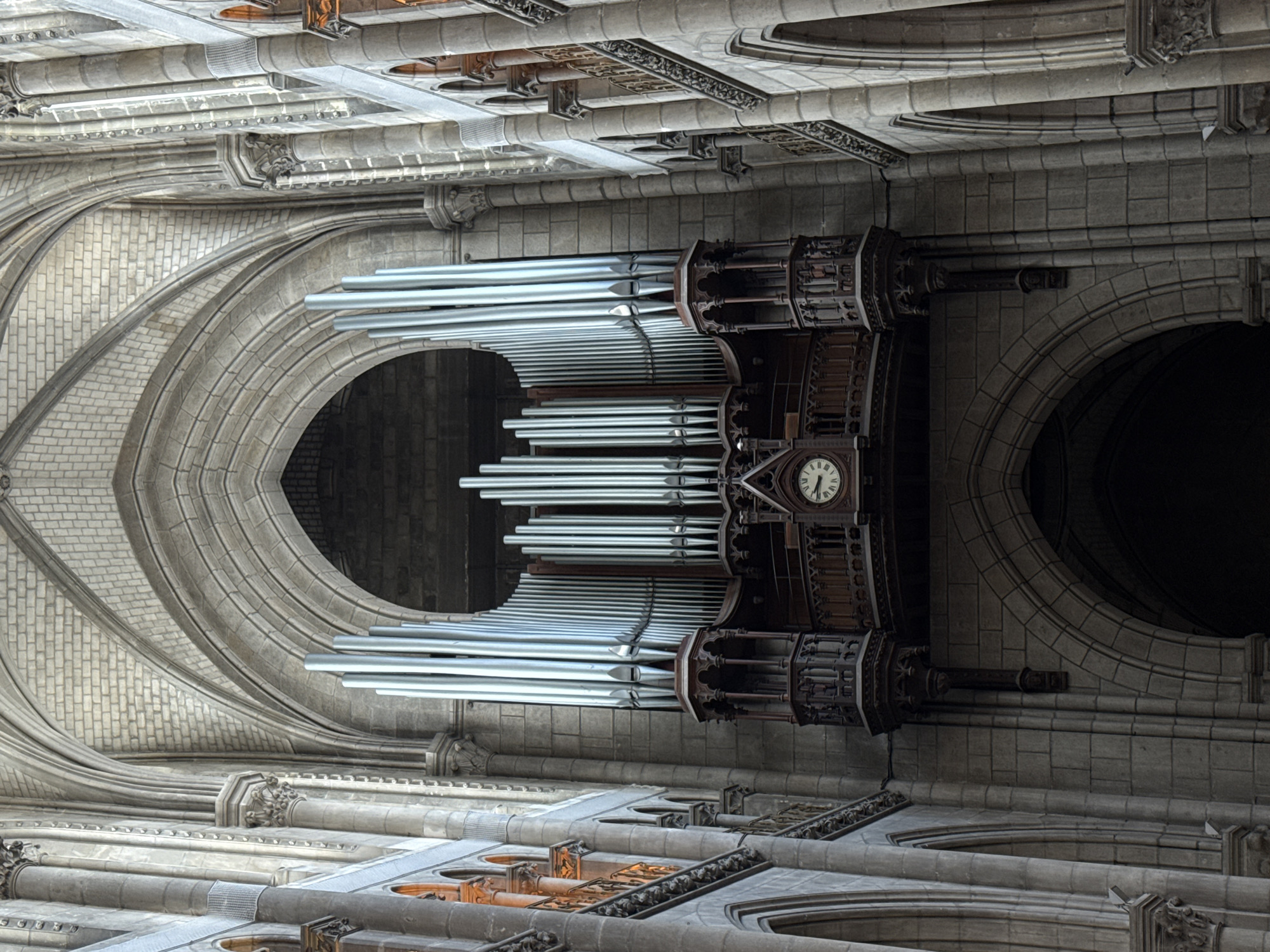 The image displays a grand pipe organ inside the Cathédrale Saint-Pierre-et-Saint-Paul de Nantes, located in Nantes, France. The organ, a prominent feature, is mounted high on a stone wall and is composed of numerous polished metal pipes arranged in multiple sections. The pipes vary in size and are predominantly silver-toned. Beneath the organ, a dark wooden structure supports it, featuring intricate carved details and balconies. A visible clock face with Roman numerals is integrated into this wooden support.
The cathedral's interior is characterized by soaring Gothic architecture, with stone arches, ribbed vaults, and large stone pillars creating a sense of height and grandeur. The walls are constructed of stone blocks, with some areas showing a finer, brick-like stonework. Natural light, hinted at by the warm glow from arched windows on the left, illuminates parts of the structure. The scene is static, with no people or immediate activity depicted, focusing solely on the architectural magnificence and the imposing organ. The environment is one of solemnity and historical significance, typical of a large cathedral.