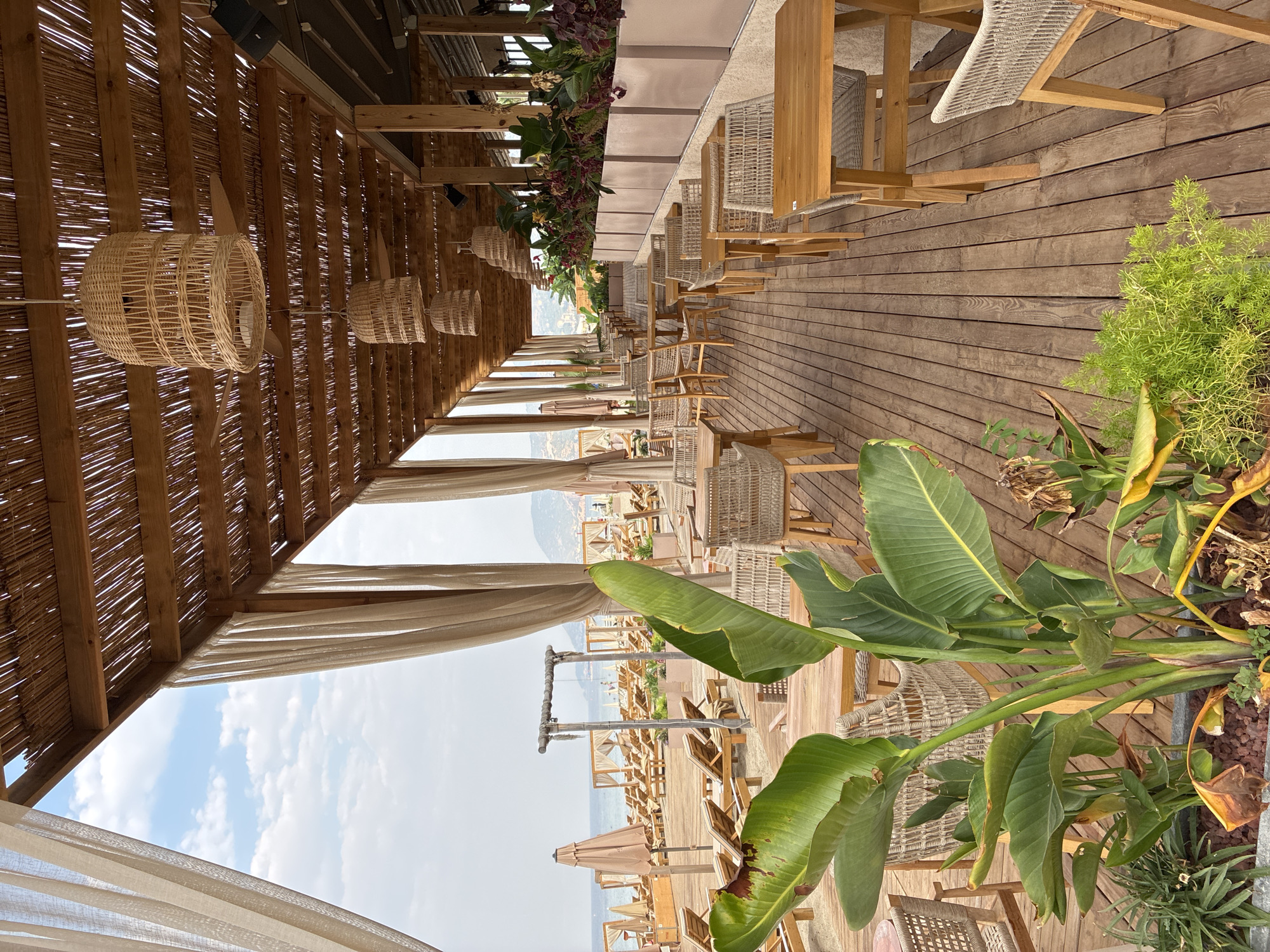 The image depicts an outdoor seating area of a beachside establishment, likely a restaurant or cafe, located in Alanya, Turkey. The setting features a wooden deck furnished with multiple tables and chairs made of woven material and wood. Overhead, a thatched roof supported by wooden beams provides shade, with woven lamps hanging at intervals. Sheer curtains drape from the roof structure along the sides. In the foreground, a large potted plant with broad green leaves adds a tropical element to the scene. Beyond the seating area, beach chairs and umbrellas are visible on a sandy beach, with the sea and a distant coastline under a partly cloudy sky. The ambiance suggests a relaxed, resort-like environment.