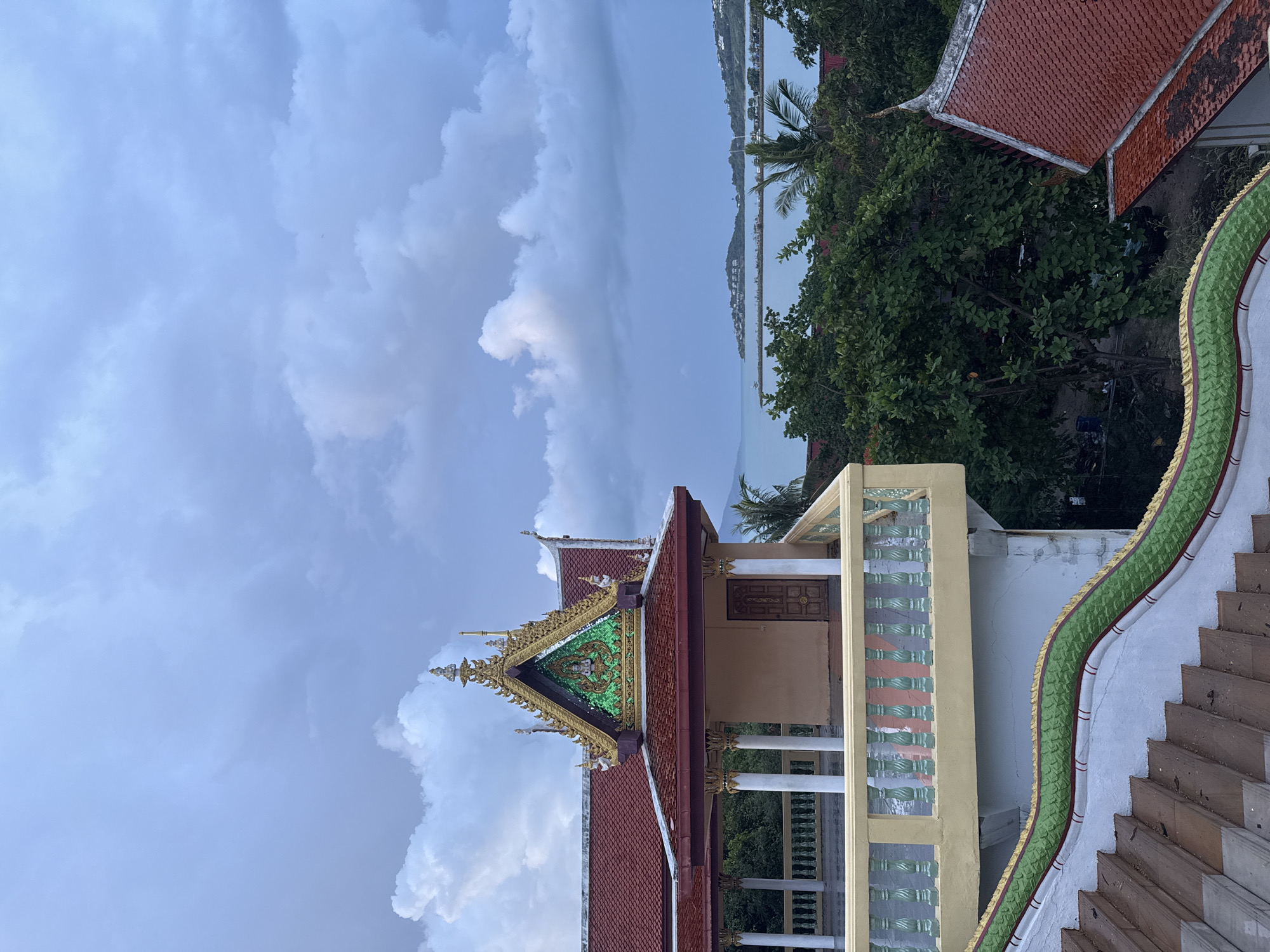 The image depicts a temple structure with a prominent red tiled roof and ornate golden decorations. The temple features a balcony with a decorative railing and a staircase adorned with a green serpent-like railing. In the background, lush green trees surround the temple, leading to a view of the ocean and a distant coastline with buildings. The sky is filled with dramatic clouds. This scene is located at Baan Plai Laem Temple in Baan Plai Laem, Thailand. No people are visible in the image.