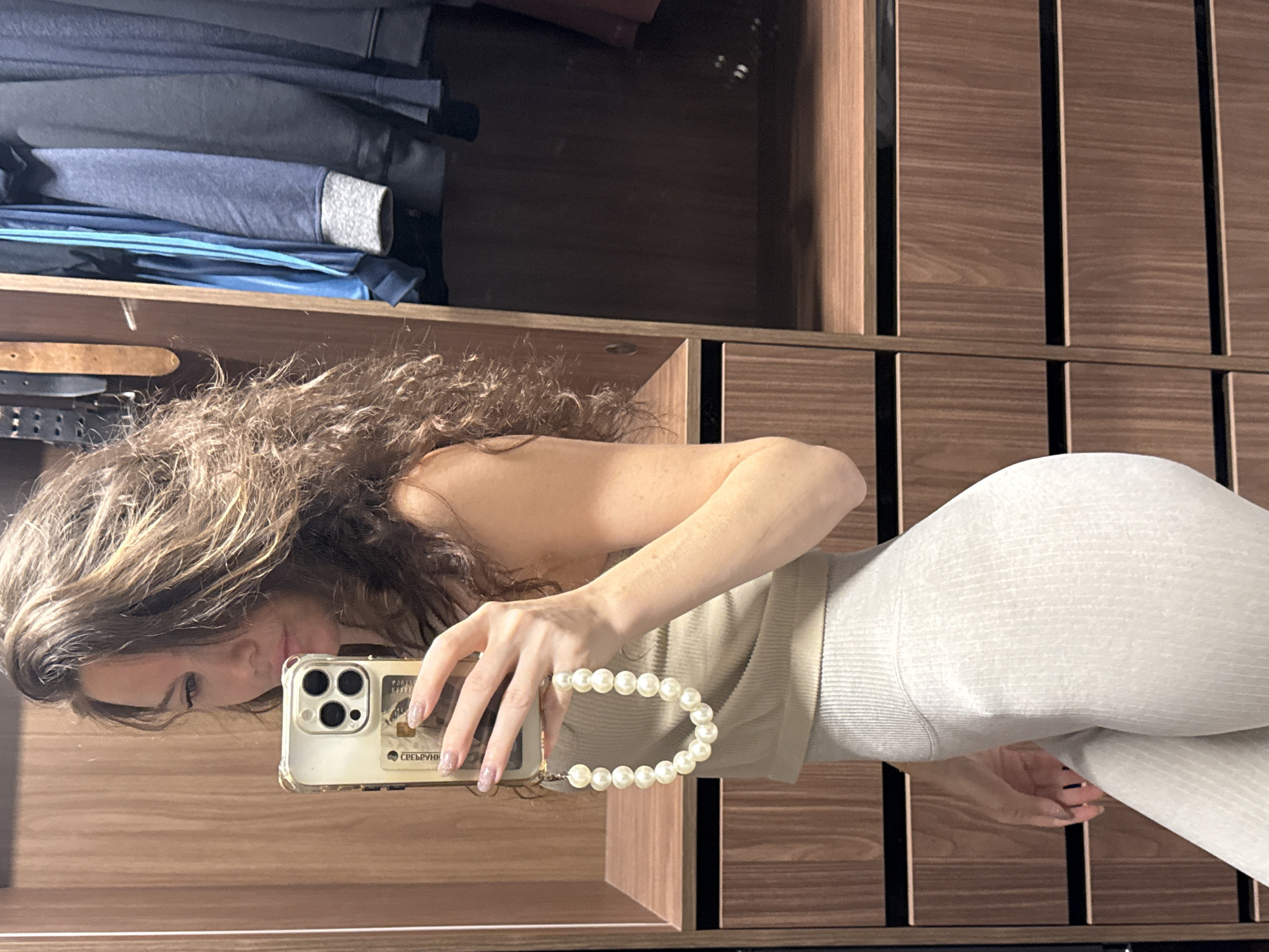 The image is a mirror selfie of a woman, oriented 90 degrees clockwise. The woman is horizontally positioned with her head on the left, looking into the camera with a slight, relaxed smile. She has long, curly dark brown hair and is wearing a beige ribbed tank top and light grey ribbed leggings. Her right hand, featuring glitter-polished nails, holds a gold-colored iPhone in a clear case, which has a white pearl wrist strap attached. Visible through the phone case is what appears to be a bank card with partially visible Russian text, "СРЕДБРУНК" (likely part of "Сбербанк"), and the year "2019".
The setting is an indoor space, likely a closet or dressing room, reflected in a mirror. The background shows dark brown wooden shelving units with vertical dividers. On an upper shelf, neatly folded clothes in blue, grey, and dark tones are stacked. Below this, a shelf contains several belts, including a prominent brown leather one. The scene is illuminated by indoor lighting. The context provided indicates the location is Moscow, Russia.