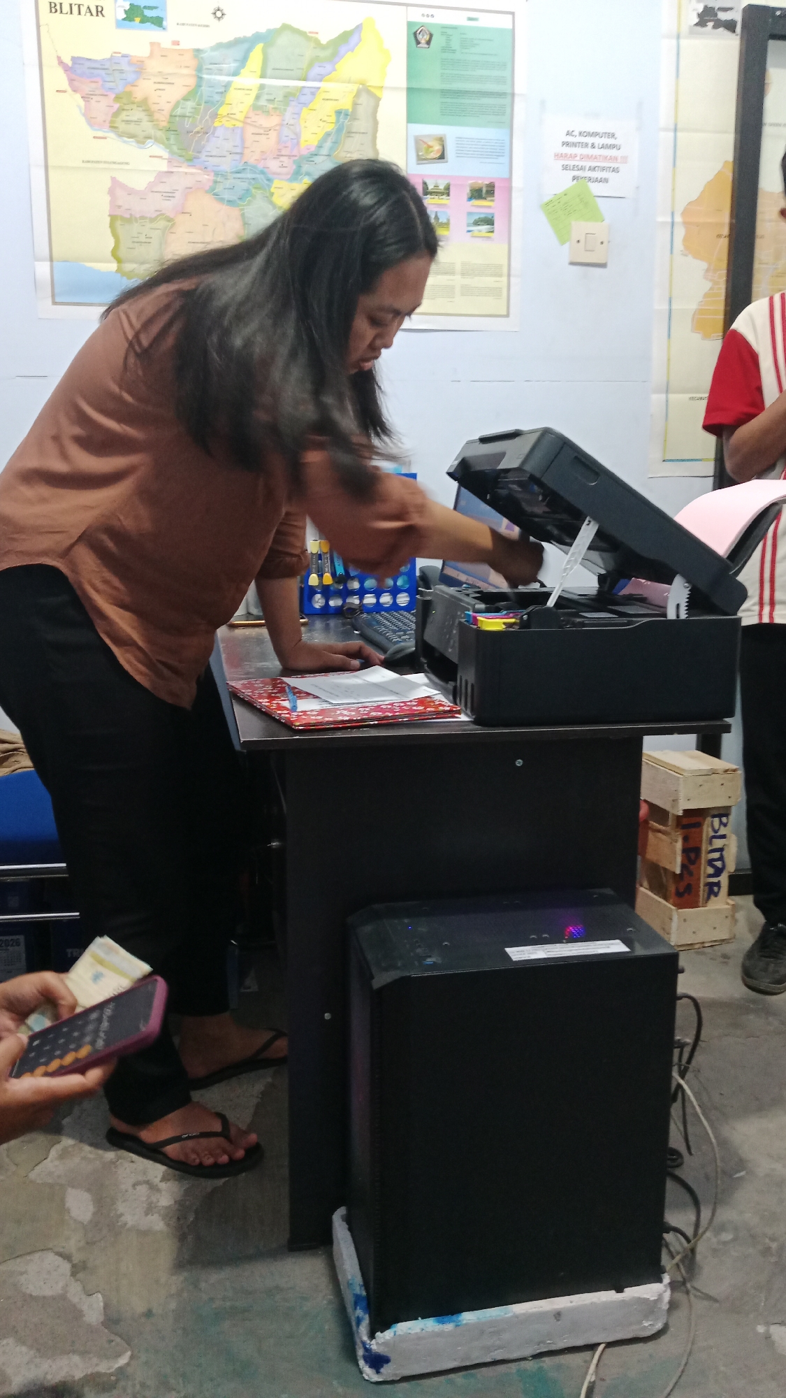 The image depicts an indoor scene likely within an office or administrative building in Blitar, Indonesia. A person with long dark hair, wearing a brown shirt and black pants, is leaning over a desk and operating a printer. The desk is dark colored and cluttered with various items including papers, a keyboard, and what appears to be a calculator on a phone held by a hand in the foreground. Behind the desk, a large black computer tower sits on a white base. The walls are decorated with maps and informational posters. One poster, located to the right of the printer, has text in Indonesian that translates to "AC, COMPUTER, PRINTER & LAMP. PLEASE SAVE ON Electricity. FINISH WORK ACTIVITIES." The floor is concrete and shows signs of wear. Another person, partially visible on the right, is wearing a red and white striped shirt and dark pants. The overall impression is of a functional workspace in operation.