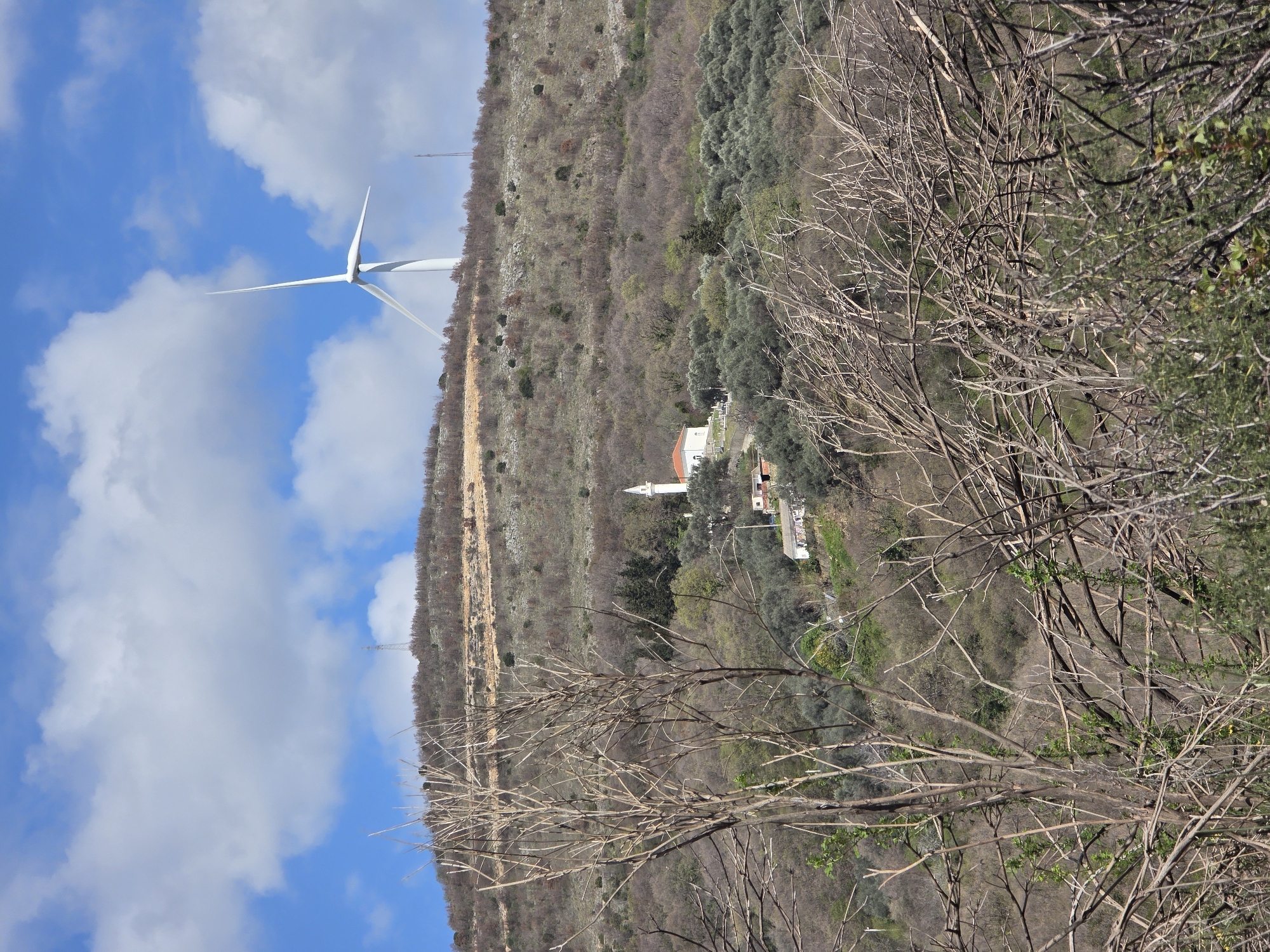 The image shows a hillside in Krute Ulcinjske - Krytha e Ulqinit, Montenegro. A wind turbine is positioned on the crest of the hill against a blue sky with scattered clouds. Lower down the hillside is a mosque with a white minaret. The foreground features bare branches of trees. The slope is vegetated with scrub and olive trees. A dirt track or road is visible winding up the hillside.