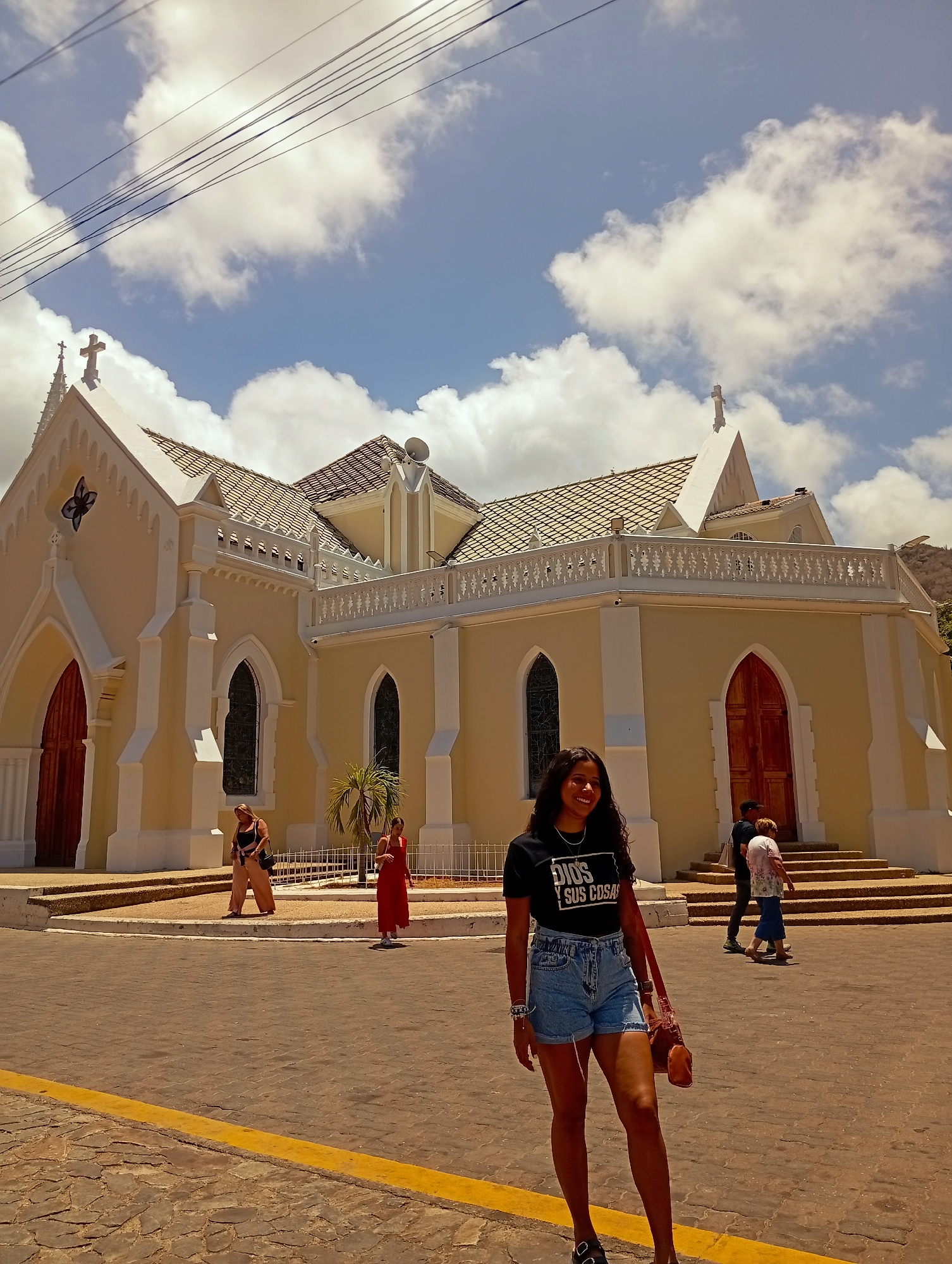 The image shows a woman standing on a brick paved street in El Valle del Espíritu Santo, Venezuela, with a church in the background. The church is light yellow with white trim, featuring pointed arches, stained glass windows, and a terracotta tile roof. Several other people are walking in front of the church. The sky is blue with scattered white clouds. Overhead power lines are visible in the top left corner. The woman in the foreground is wearing denim shorts and a black t-shirt.