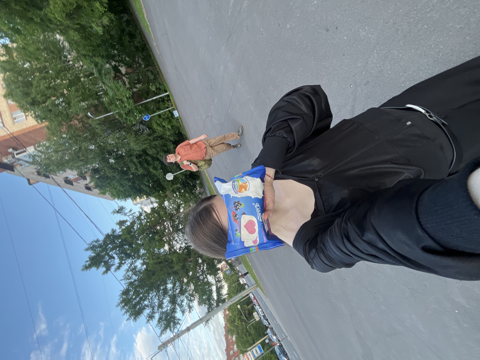 The image captures an outdoor scene, likely a street or wide paved path, in what appears to be an urban or suburban setting in Saint Petersburg, Russia.
In the foreground, a person is prominently featured, holding a blue package in front of their face, partially obscuring it. They are wearing a black long-sleeved top or jacket, and black trousers with a light-colored belt buckle visible. A red string or bracelet is on their right wrist. The blue package features cartoon graphics, including a cow and different ice cream bars, notably a heart-shaped one and another with berries. Visible text on the package includes "Пломбир" (Plombir, a type of ice cream) and "Коровка из Кореновки" (Korovka iz Korenovki), a recognizable Russian brand.
Further in the background, another person is walking away from the camera on the paved surface, looking slightly over their shoulder. This individual is dressed in an orange-brown long-sleeved shirt, light brown trousers, and carries a green backpack.
The setting includes a paved dark gray surface, abundant green trees lining the side, and multistory buildings visible behind the foliage. Overhead wires traverse the sky, and streetlights and a blue circular road sign are discernible along the path. The sky is clear and bright blue with some scattered white clouds, indicating a daytime scene, possibly late afternoon or early evening. The overall atmosphere is casual and relaxed.