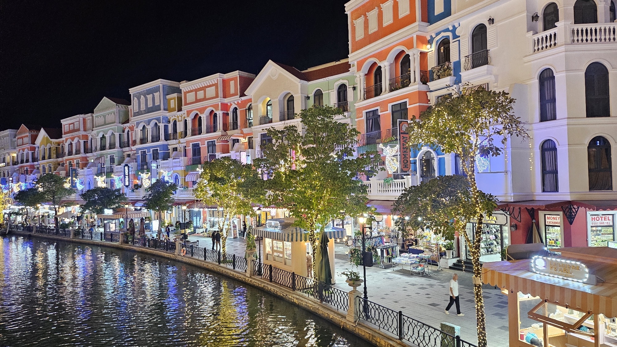 This is a nighttime photograph of the Sunset Town in Phú Quốc, Vietnam, featuring a canal lined with colorful buildings. The architecture is European-inspired with pastel facades, arched windows, and balconies. Trees adorned with fairy lights are scattered along the canal, reflecting in the water. Several small kiosks and shops are visible along the pedestrian walkway, with some displaying goods and signage. People are seen walking along the path and interacting with the shops. The overall atmosphere is festive and vibrant, illuminated by the building lights and string lights.