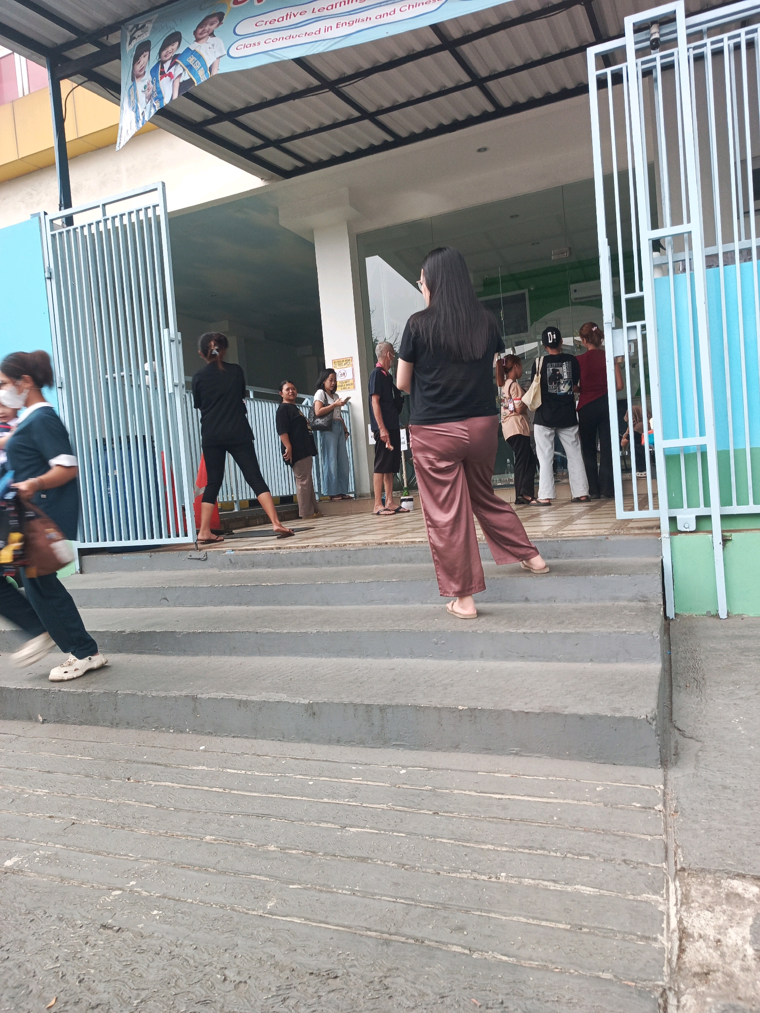 In Pakulonan Barat, Indonesia, a group of people stand on the steps outside a building entrance. The building features a light blue and green facade with a white metal gate. A banner with images of children and text reading "Creative Learning" and "Class Conducted in English and Chinese" is displayed above the entrance. A woman in a dark blue uniform and mask walks down the steps from the left, carrying a brown bag. Another woman in a black top and pink pants stands on the steps facing away from the camera. Several other individuals are visible standing or walking around the entrance. The ground is paved with concrete.