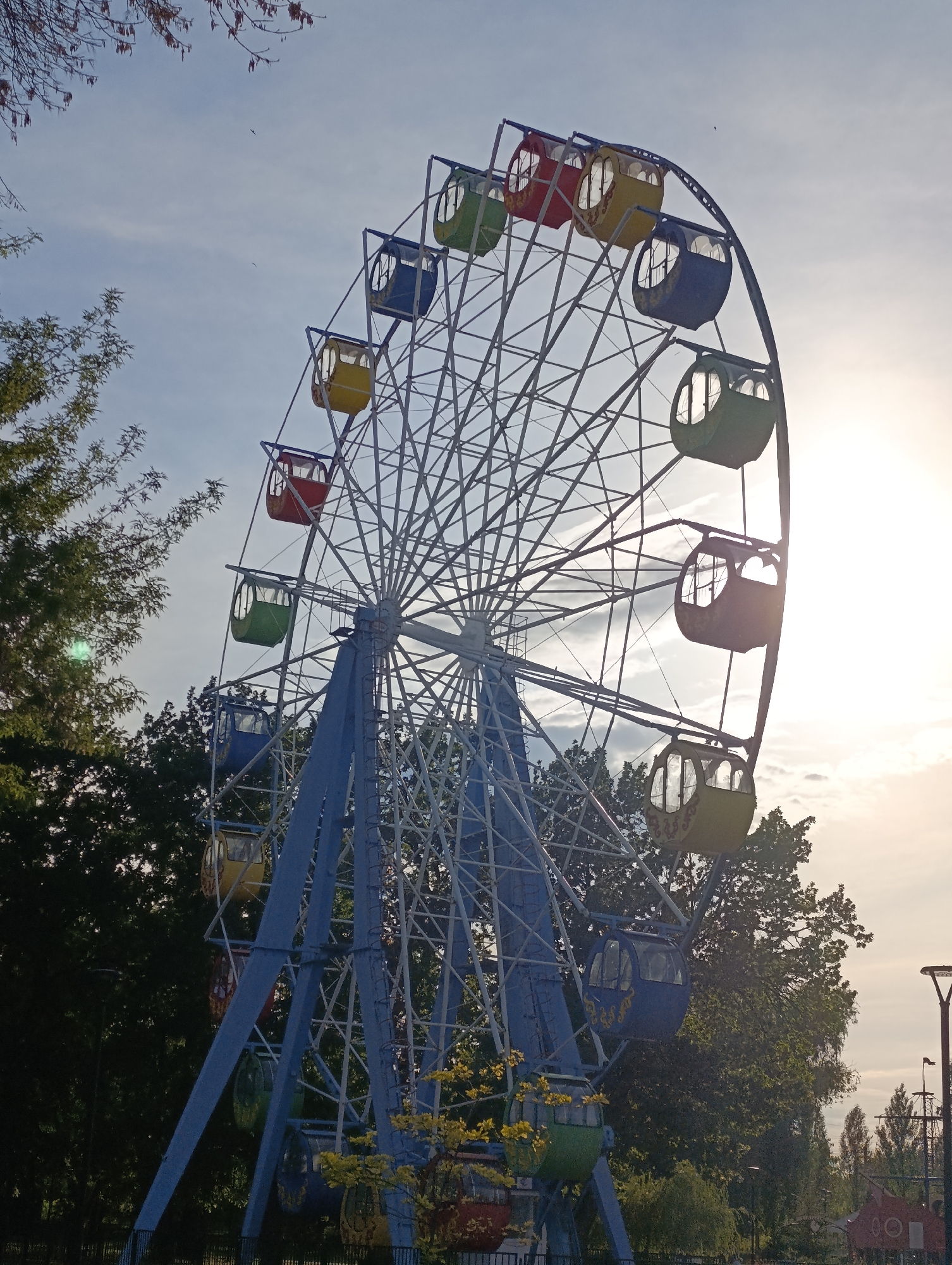 A large Ferris wheel with colorful gondolas stands tall against a bright, slightly hazy sky, suggesting late afternoon or early morning light. The sun is visible, casting a lens flare and illuminating the sky with a warm glow. The Ferris wheel structure is painted blue, with a white metal frame supporting the circular arrangement of gondolas, each a distinct color: red, yellow, green, and blue. Some gondolas appear weathered, hinting at the age of the amusement.
The Ferris wheel is situated in a park-like setting, surrounded by lush green trees with some branches reaching into the frame, particularly from the upper left. The lower part of the image shows more dense foliage and hints of park amenities, including a dark metal fence and what appear to be decorative planters. In the distance, a lamppost and silhouettes of other amusement park structures, including what might be a small sailboat ride, are visible. The overall scene evokes a sense of quietude and nostalgia, perhaps a break in the day's activities at an amusement park. The image was taken in Novokuybyshevsk, Russia. There is no visible text.