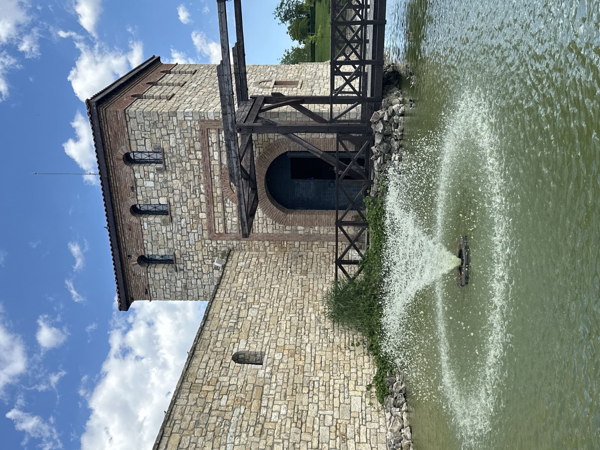 The image depicts a historical structure, likely a fortress or castle, in Neofit Rilski, Bulgaria. The building is constructed of stone and brick, with arched windows and a large arched entrance. A wooden drawbridge extends from the structure over a body of water, where a fountain is actively spraying water. The water is green, suggesting it might be a pond or a lake. The sky is blue with scattered white clouds. There is a wooden fence along the edge of the water. The overall scene evokes a sense of history and tranquility, with the natural elements of water and sky complementing the ancient architecture. No people are visible in the image.