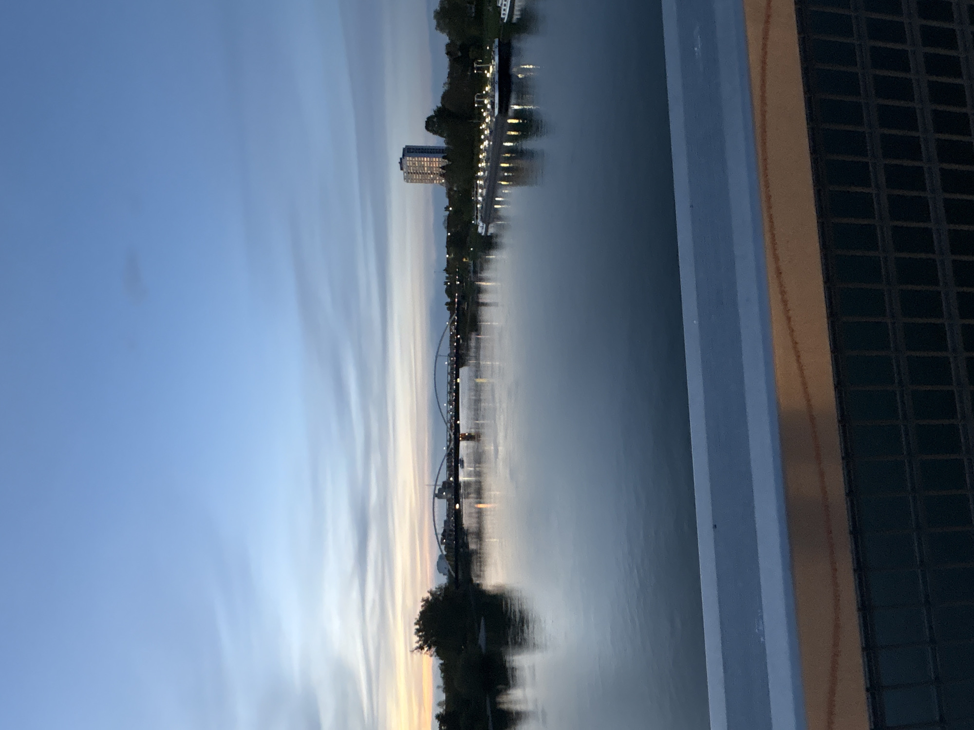 The image presents a serene twilight view of a city riverfront, likely in Strasbourg, France, captured from an elevated vantage point. The sky, a gradient of soft blue and grey, shows subtle streaked cloud patterns, indicating dusk. Below, a wide body of water, possibly the Ill River, acts as a reflective mirror, capturing the fading light of the sky and the warm glows of urban illumination.
Along the opposite bank, a cityscape emerges. Numerous artificial lights from buildings and streetlamps are visible, casting elongated, shimmering reflections on the calm water. A prominent, multi-story building with many lit windows stands out among the structures. Further along the river, an elegant arch bridge spans the water, its form delineated by lights that also reflect on the surface below. Trees and foliage line the banks, appearing as dark silhouettes against the ambient light of the city.
In the immediate foreground on the right, a section of a building or structure is visible, featuring a pale blue vertical element alongside a beige wall with a dark grid pattern, possibly part of a window or balcony from where the photo was taken. No discernible activity or people are present. The overall atmosphere is peaceful and contemplative, showcasing the urban landscape bathed in the soft glow of evening. No visible text is present in the scene.