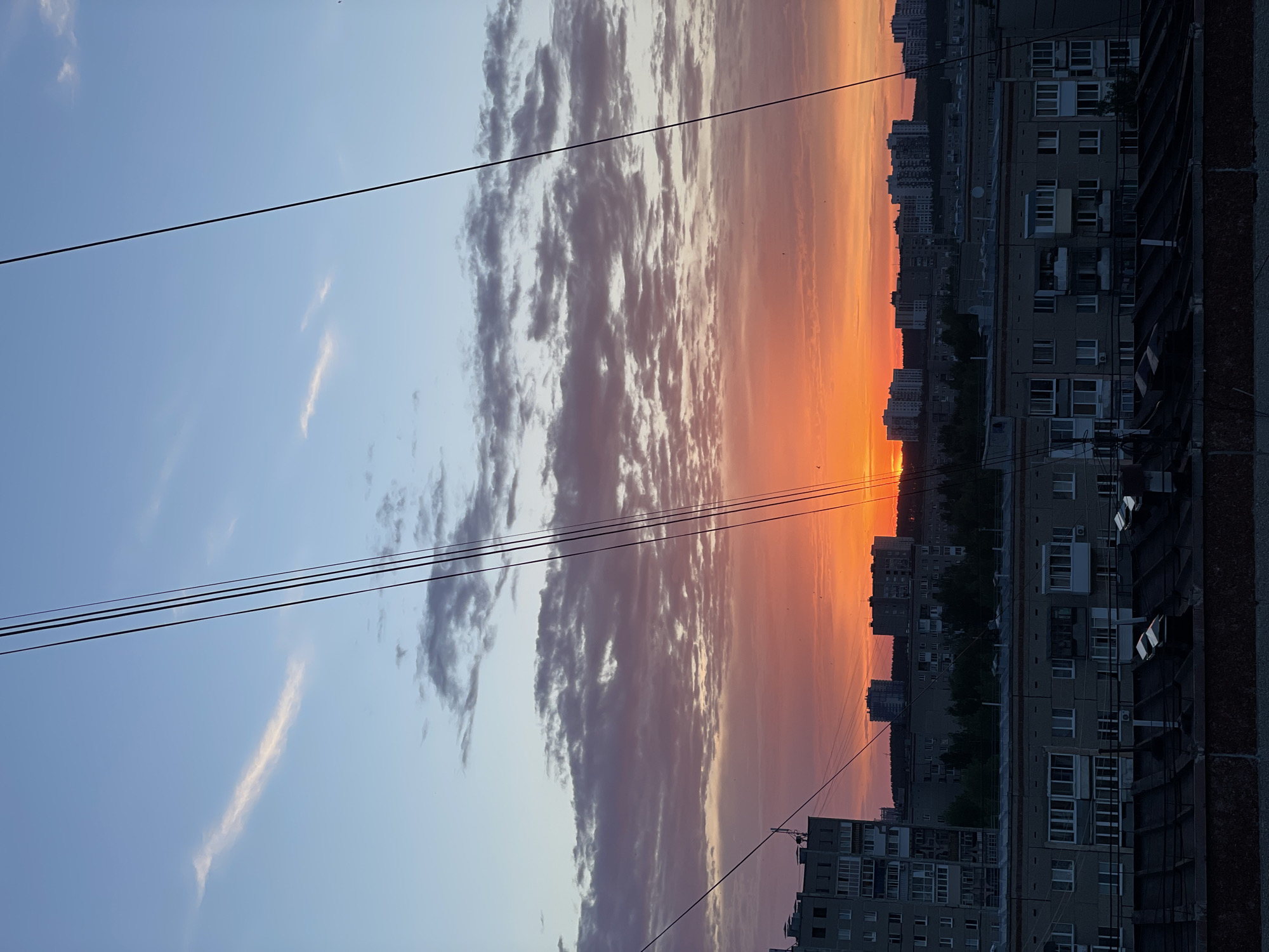 The image shows a cityscape at sunset, likely in Izhevsk, Russia, though precise confirmation would require local knowledge or comparison with specific landmarks. The sky transitions from blue to orange, indicating the sun is near the horizon. Clouds are present, with some near the horizon appearing dark. Power lines stretch across the sky in the foreground. In the background, there are several multistory residential buildings. The buildings are typical of urban architecture in former Soviet countries, with multiple rectangular windows. Vegetation, likely trees, is visible between the buildings. A dark wall, possibly a balcony or another part of a building, is present in the right foreground. There are no people visible in the image.