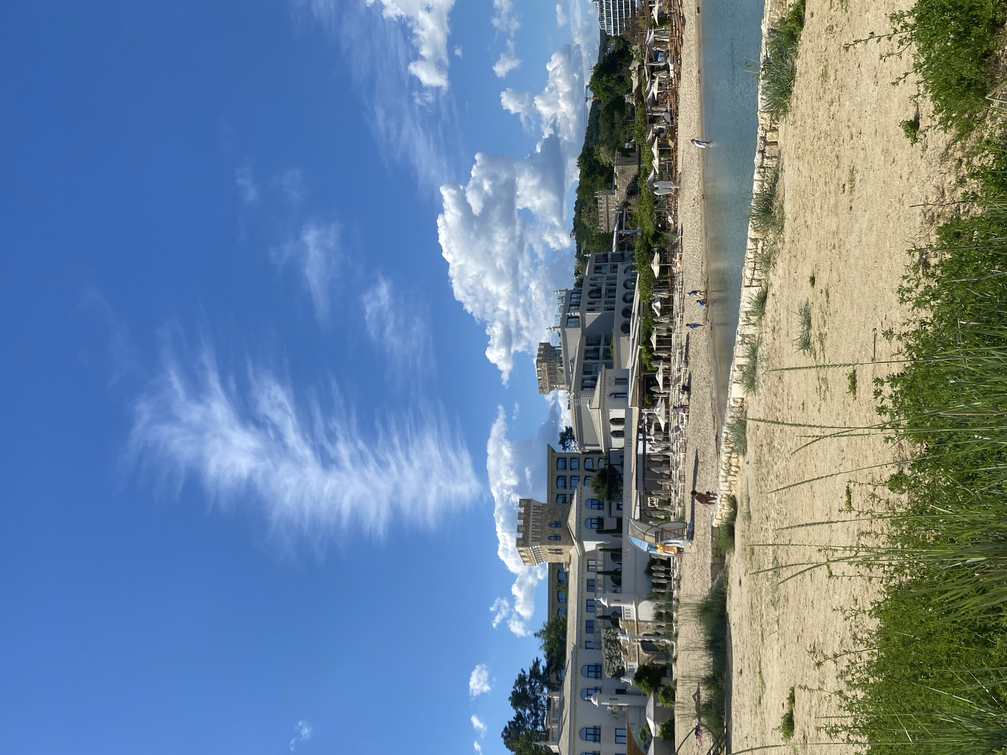 The image shows a view of the beach, buildings, and sky in Varna, Bulgaria. The sky is blue with cirrus and cumulus clouds. The beach is sandy and runs parallel to buildings. The buildings are modern, white and appear to be residential and/or tourist accomodations. Beach chairs and umbrellas are visible on the beach. There are people in the scene. The overall environment appears to be sunny and calm. Green vegetation grows near the edge of the beach.