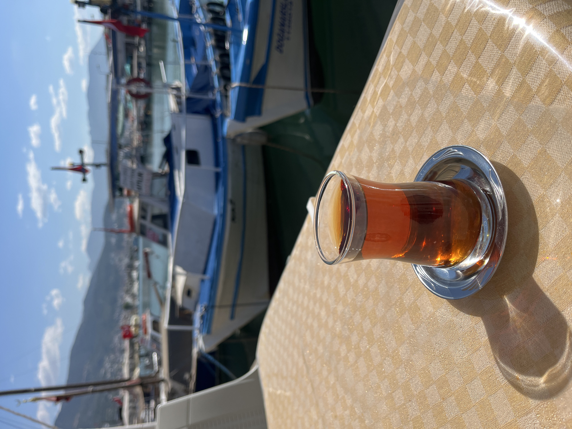A cup of Turkish tea, filled with amber liquid, sits on a small, ornate, silver saucer on a table covered with a light-yellow checkerboard tablecloth. In the background, slightly out of focus, are several docked boats, with blue and white hulls, and red flags. The scene is set in Alanya, Turkey, evidenced by the distinct Turkish tea glass and the presence of boats typical of a coastal town. Mountains are visible in the distant background under a bright blue sky.
