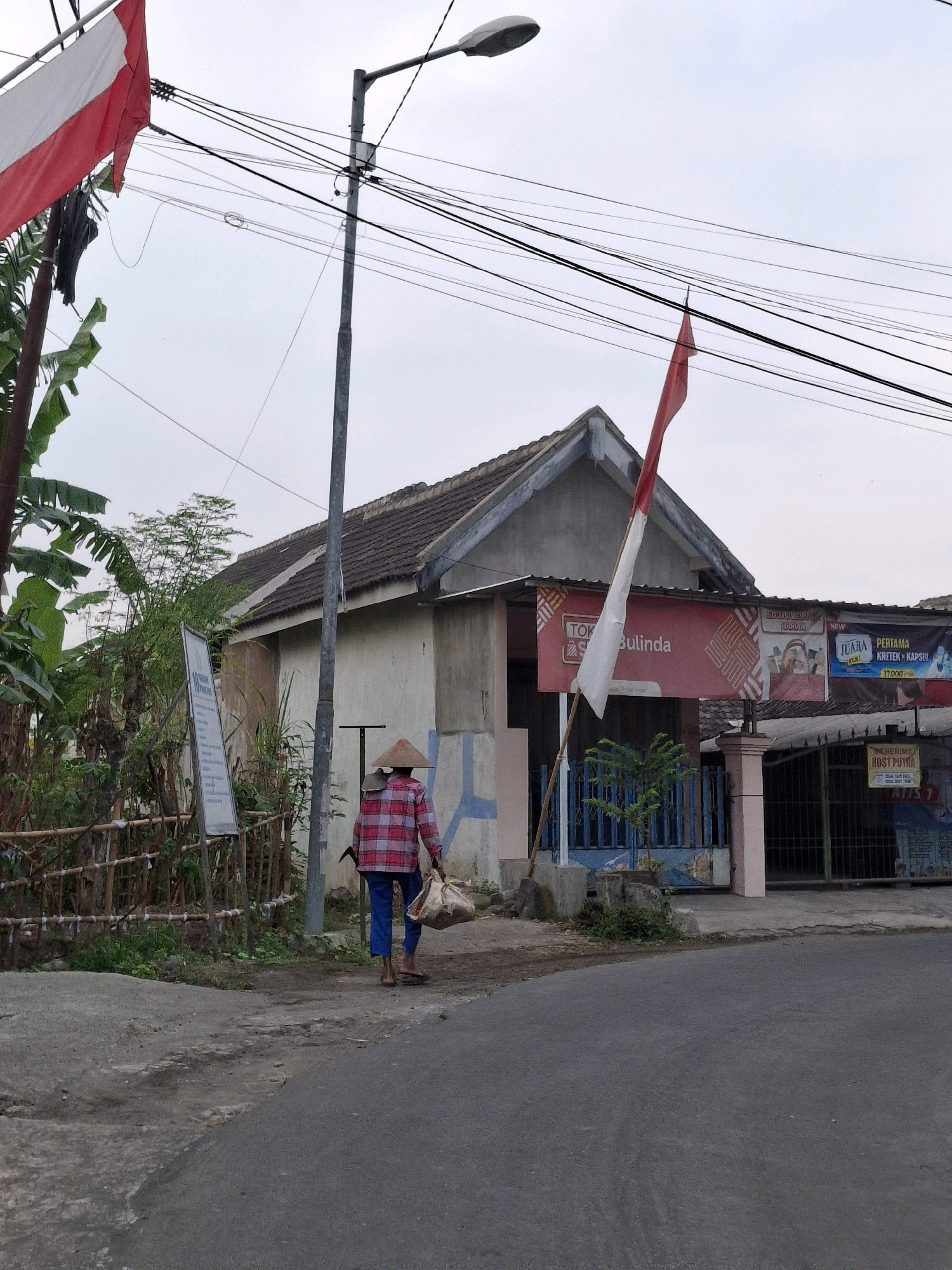 In Lumajang, Indonesia, a person wearing a traditional conical hat and a red and white checkered shirt walks away from the camera on a paved road. They are carrying two large bags, one slung over their shoulder and another in their hand. Behind them, a small building with a gray tiled roof is visible, adorned with red and white banners. A flagpole with a red and white flag stands in front of the building. The surrounding area features lush green foliage, a bamboo fence, and a street lamp. The sky is overcast.