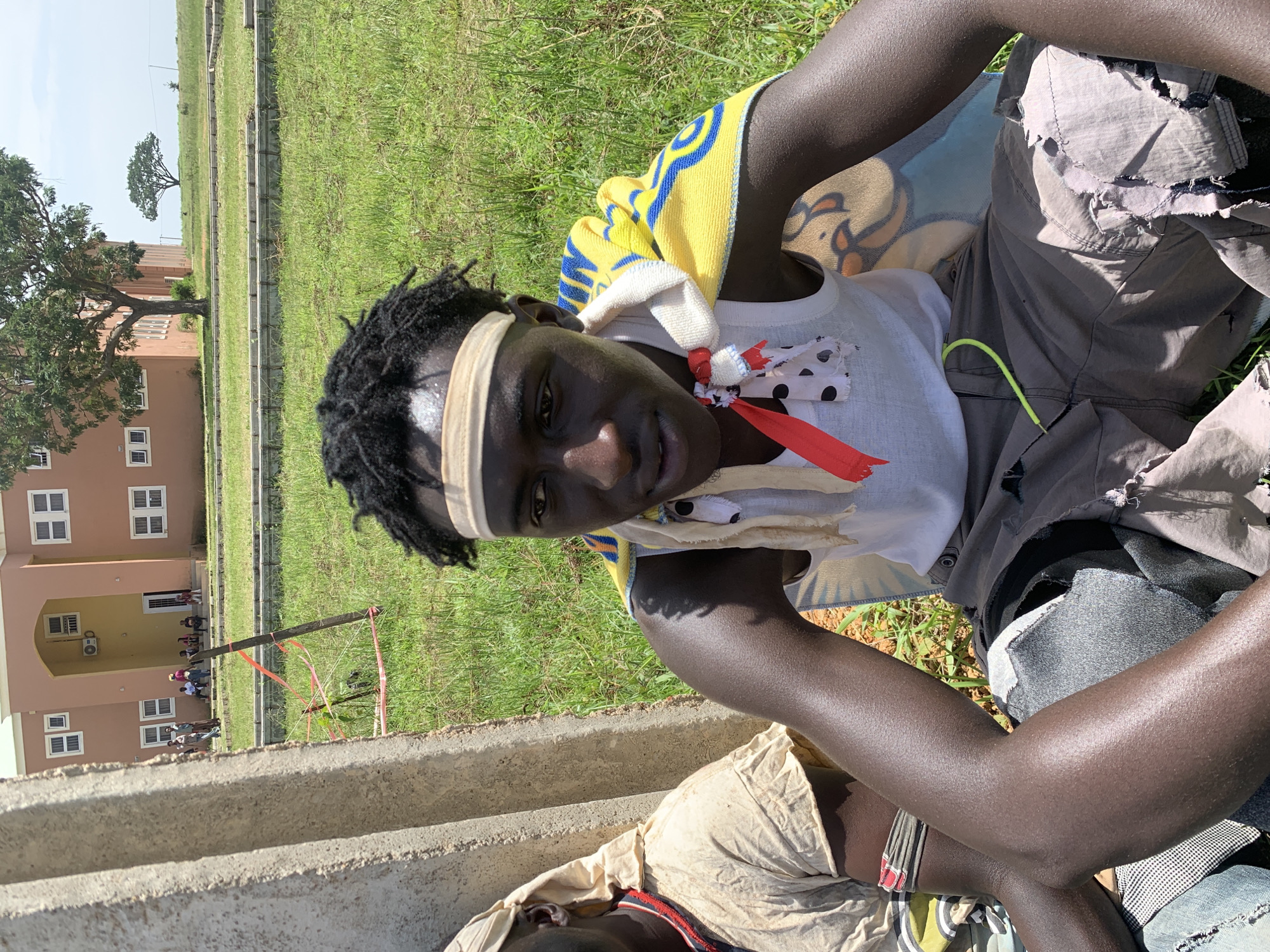 The image depicts a young Black male seated outdoors in what appears to be Gwafan, Nigeria. He is wearing a white tank top, grey shorts with distressed detailing and a green drawstring, and a yellow and blue patterned towel draped over his shoulders. A light-colored headband is visible on his forehead, and a white cloth with black polka dots and red ribbons is tied around his neck. He is positioned in the foreground, with his arms resting on his lap. To his left, another individual wearing a light-colored garment and a patterned headband is partially visible. The background features a large, light-pink building with multiple windows and air conditioning units, indicative of a public institution or residential structure. Adjacent to the building is a large tree, and beyond that, a landscape of grassy fields and some wooden structures, possibly indicating ongoing construction. The overall environment is sunny, with the ground covered in green grass and some patches of exposed earth.