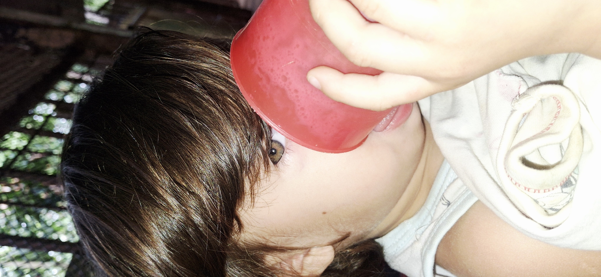 In Vallecito, Venezuela, a close-up, low-angle shot captures a young child holding a red cup to their lips, appearing to drink. The child's right eye, a hazel color, is visible, peeking out from behind the cup and a curtain of dark brown hair. The child's skin is fair, with a small mole visible on the cheek. They are wearing a light-colored sleeveless shirt, partially covered by a white cloth with red stitching. The background is blurred, suggesting an outdoor setting with foliage and trees, possibly a natural or semi-natural environment. The lighting is soft and diffused, casting a warm glow on the child's face.