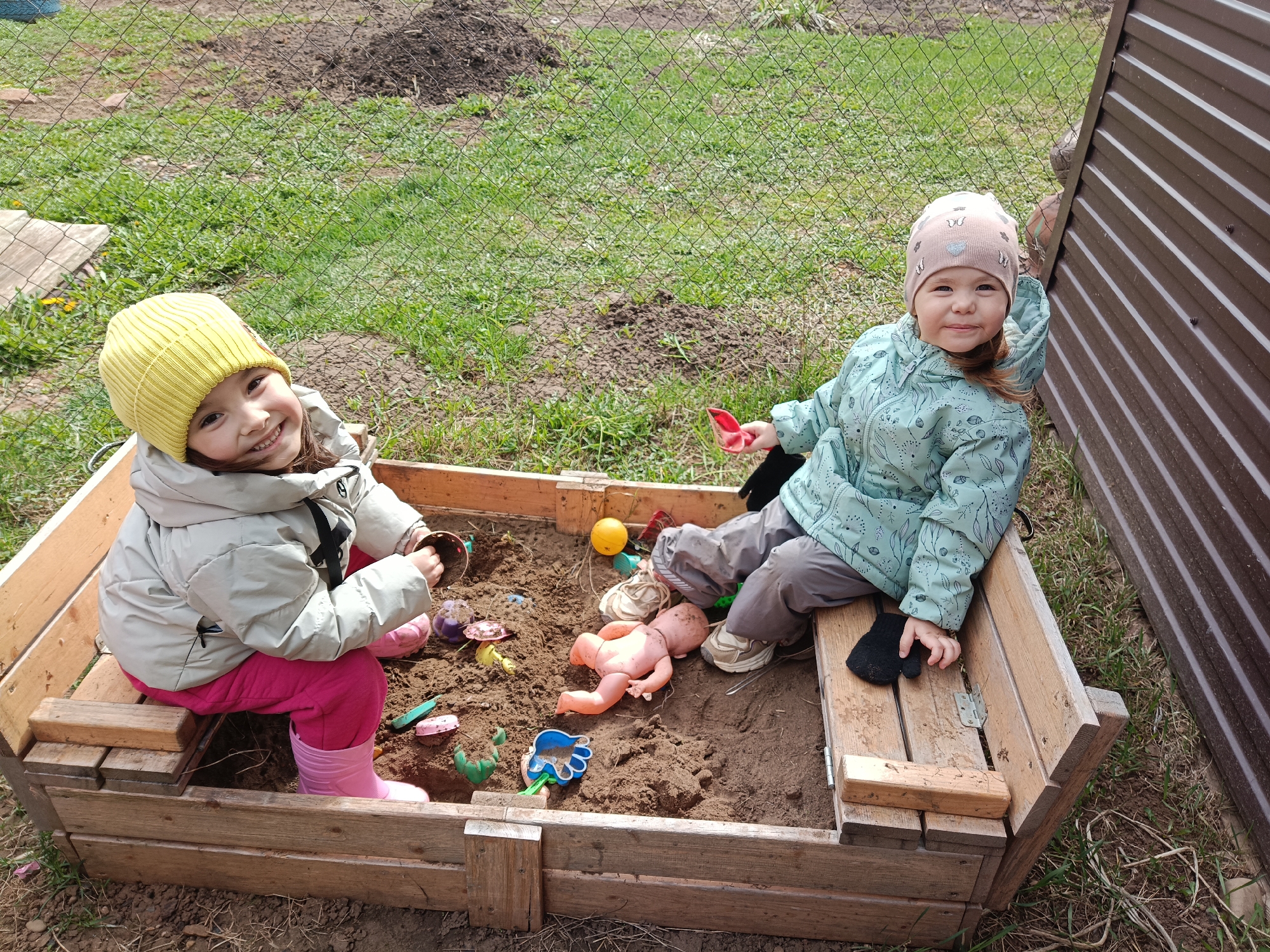 Two young girls are playing in a wooden sandbox. The girl on the left wears a yellow knit hat, a light gray jacket, pink pants, and pink boots. She is holding a toy pot. The girl on the right wears a pink hat, a turquoise jacket with a leaf pattern, and gray pants. She is holding a small red shovel in her right hand and a black glove rests on the bench behind her. The sandbox is filled with sand and various toys, including a doll, a yellow ball, and plastic molds. The background shows a green grassy area, a fence, and a brown metal structure. The location is likely a residential backyard in Barda, Russia.