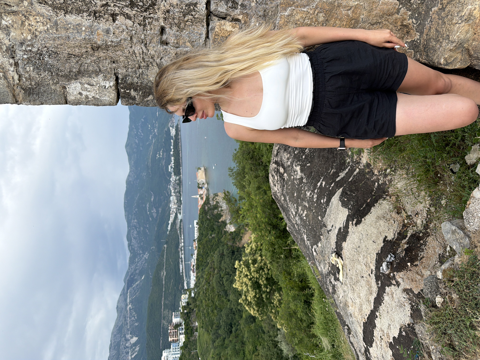 A woman with long blonde hair, wearing sunglasses, a white tank top, and black shorts, sits on a rock outcrop overlooking the city of Budva, Montenegro. The city is situated between a bay and tall, rocky mountains. The landscape includes a coastline with buildings, trees, and the Adriatic Sea. The rock formations in the foreground appear to be natural, covered with patches of grass and other vegetation. The top of the image shows a stone wall or structure. The weather appears to be overcast, with a cloudy sky visible.